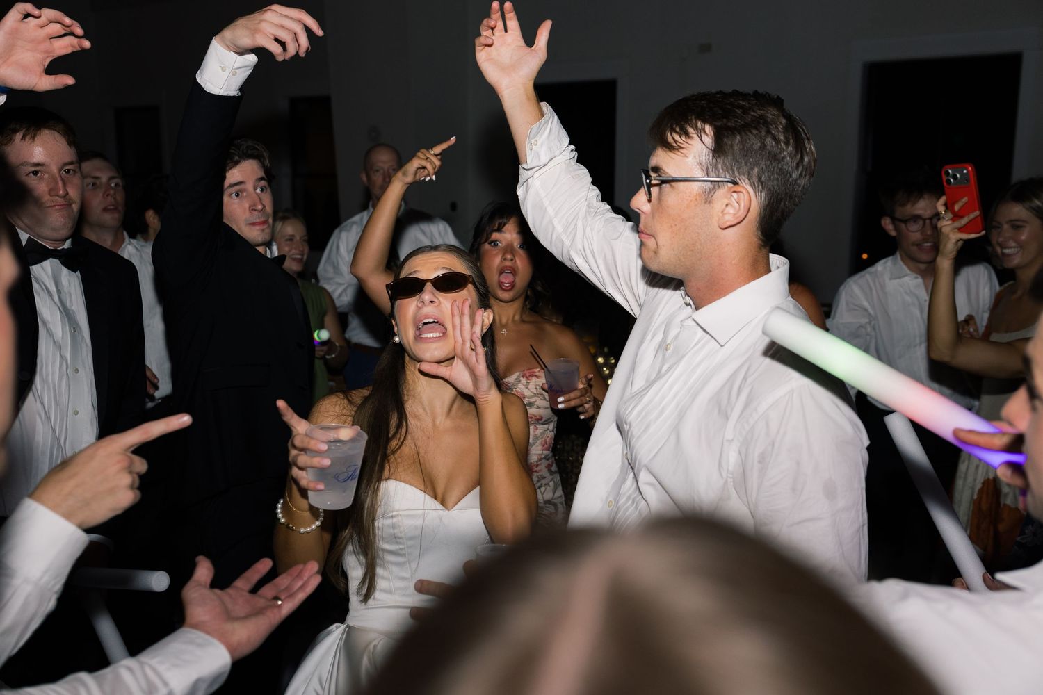 Bride and groom dancing joyfully at wedding reception party with guests cheering and glowing light sticks.