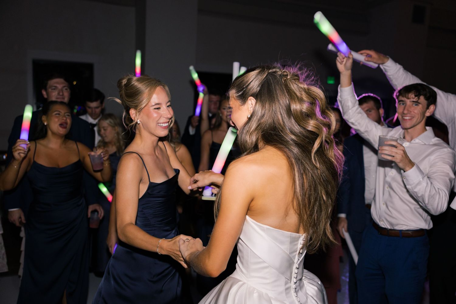 Bride in white gown dancing with guest in navy dress at wedding reception with glowing foam sticks.