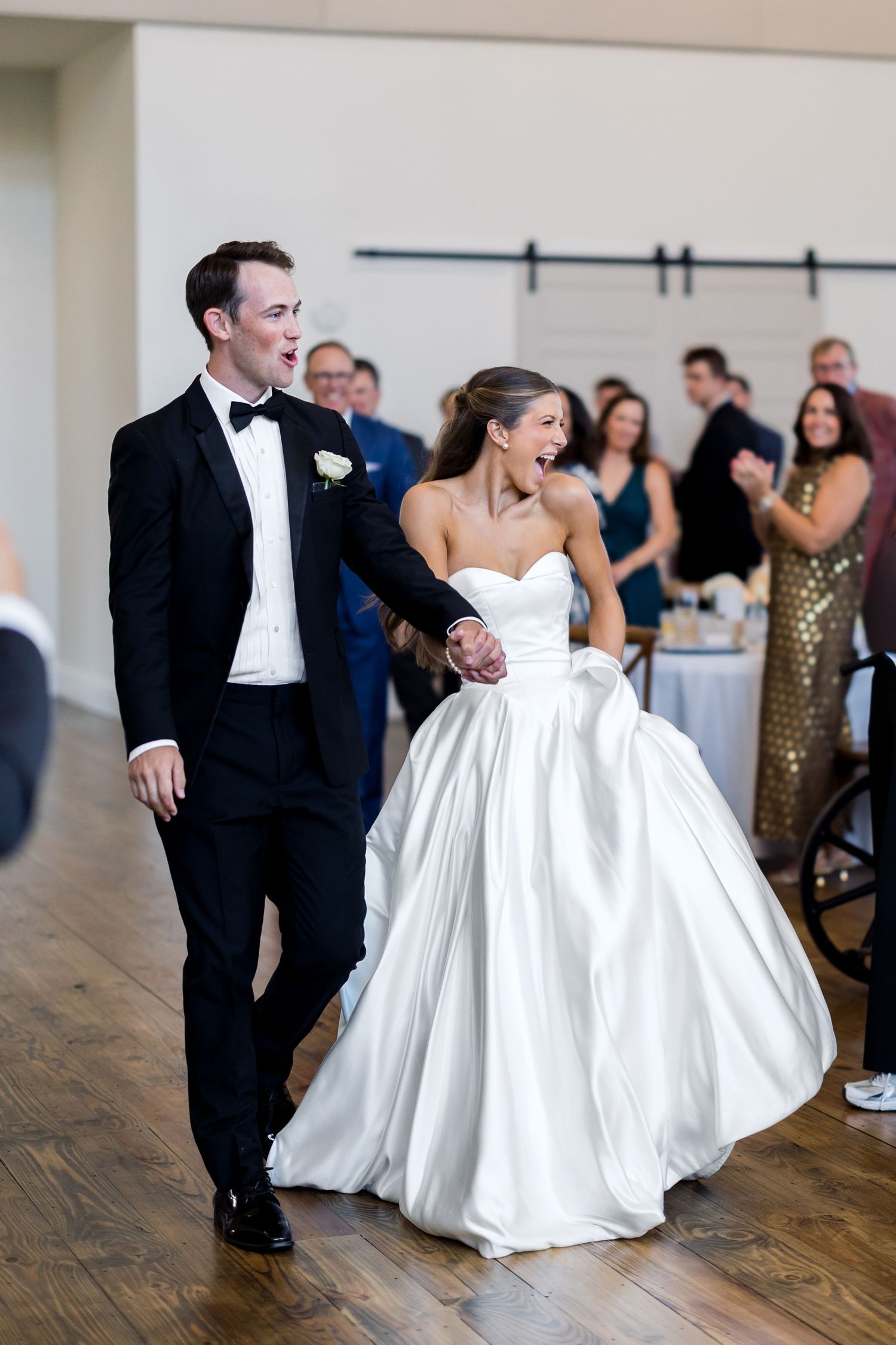 Bride and groom smile while entering wedding reception, she in a white ballgown, he in a black tuxedo.