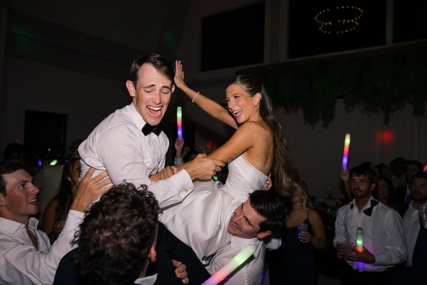 Bride and groom lifted by guests on wedding dance floor with colorful glow sticks at festive reception.