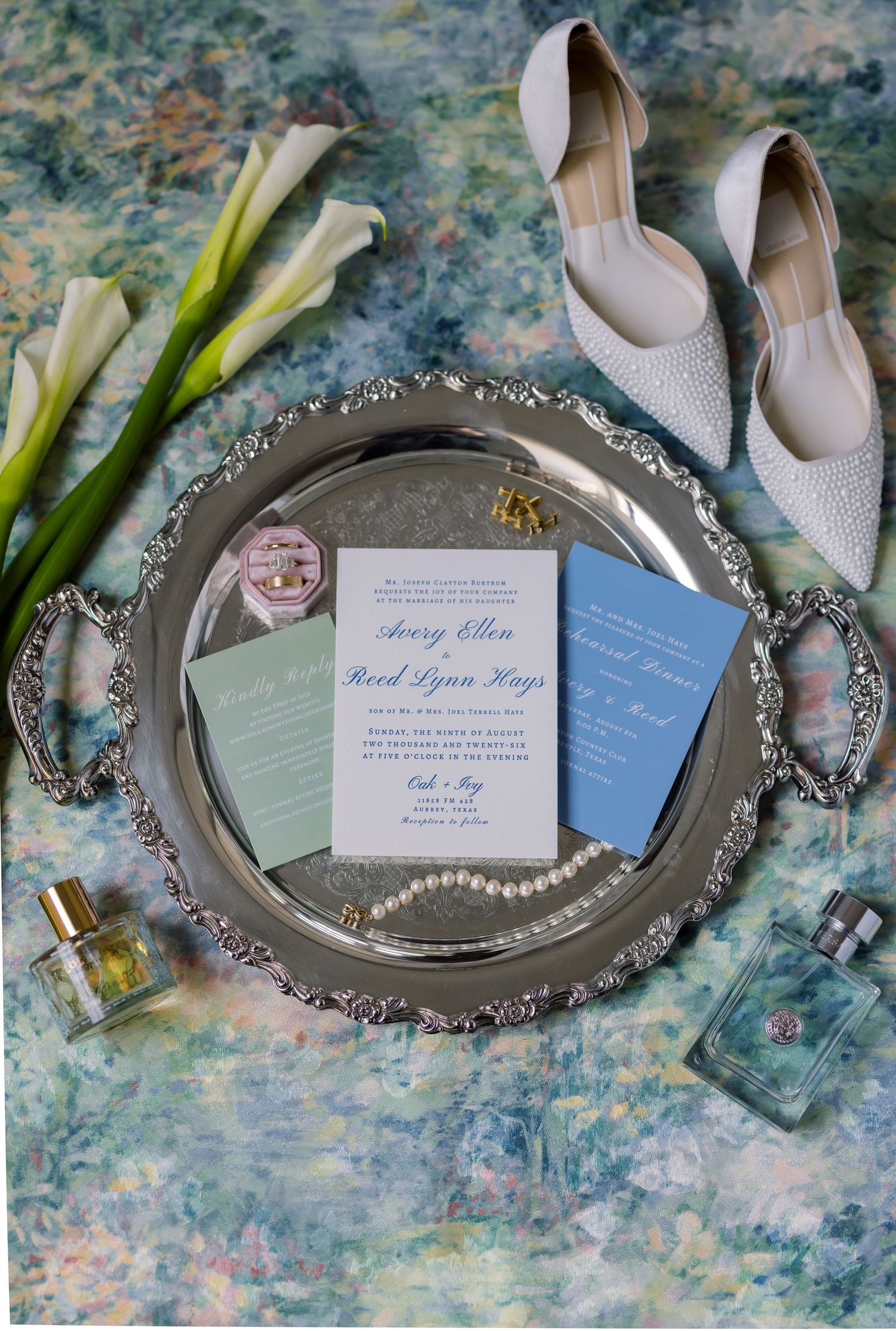 Silver tray with wedding invitations, pearl necklace, perfume bottles, white calla lilies, and bridal heels on floral backdrop.