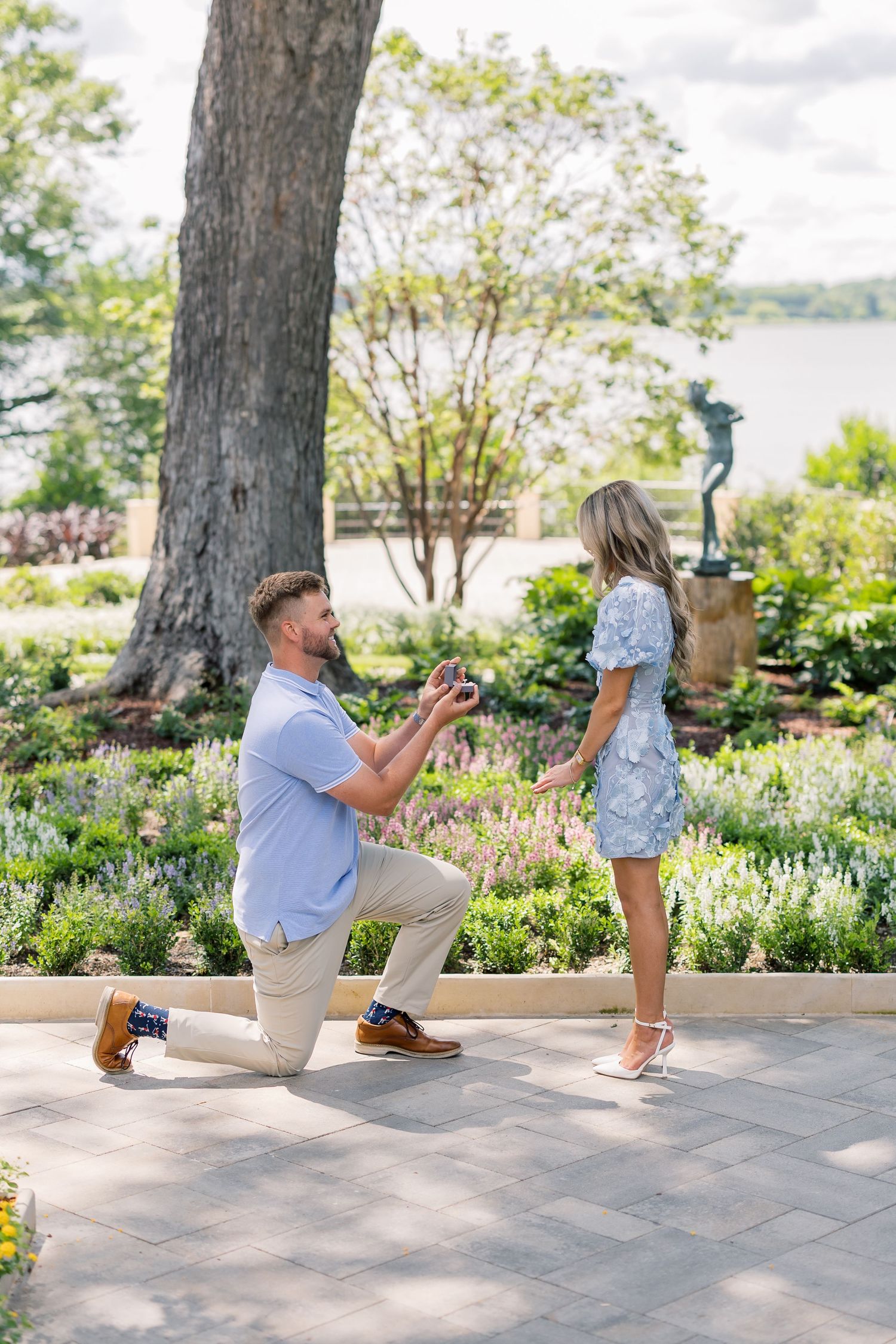 Man kneeling on one knee proposing to woman in blue dress in a lush garden with a lake in the background.