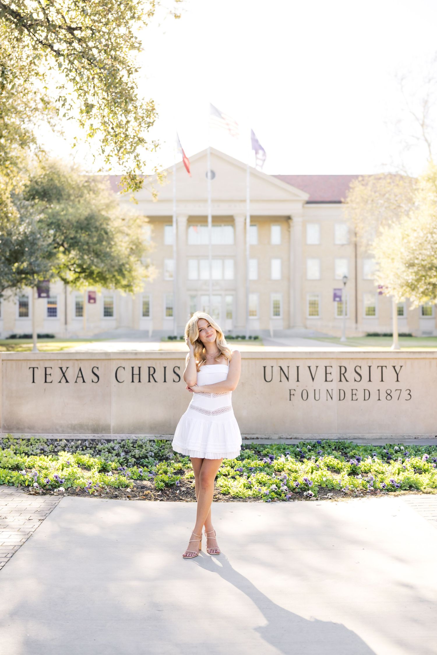 Blonde woman in white dress posing in front of Texas Christian University Founded 1873 sign.