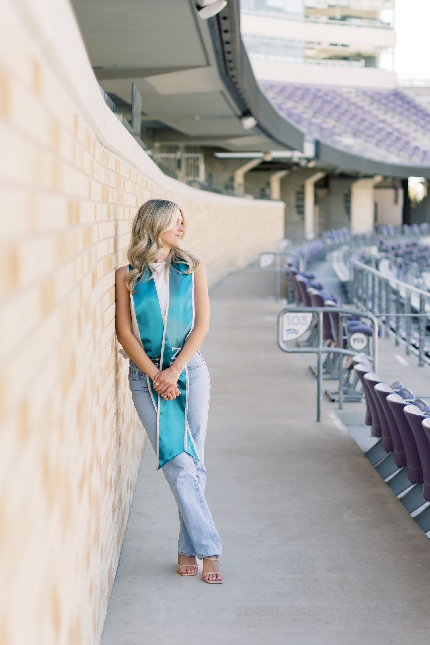 Blonde graduate in teal ZTA stole leaning against brick wall in stadium corridor.