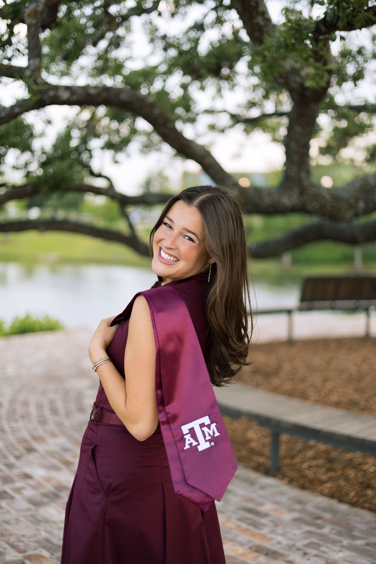 Joyful woman wearing Texas A&M maroon graduation stole posing on brick path near pond.