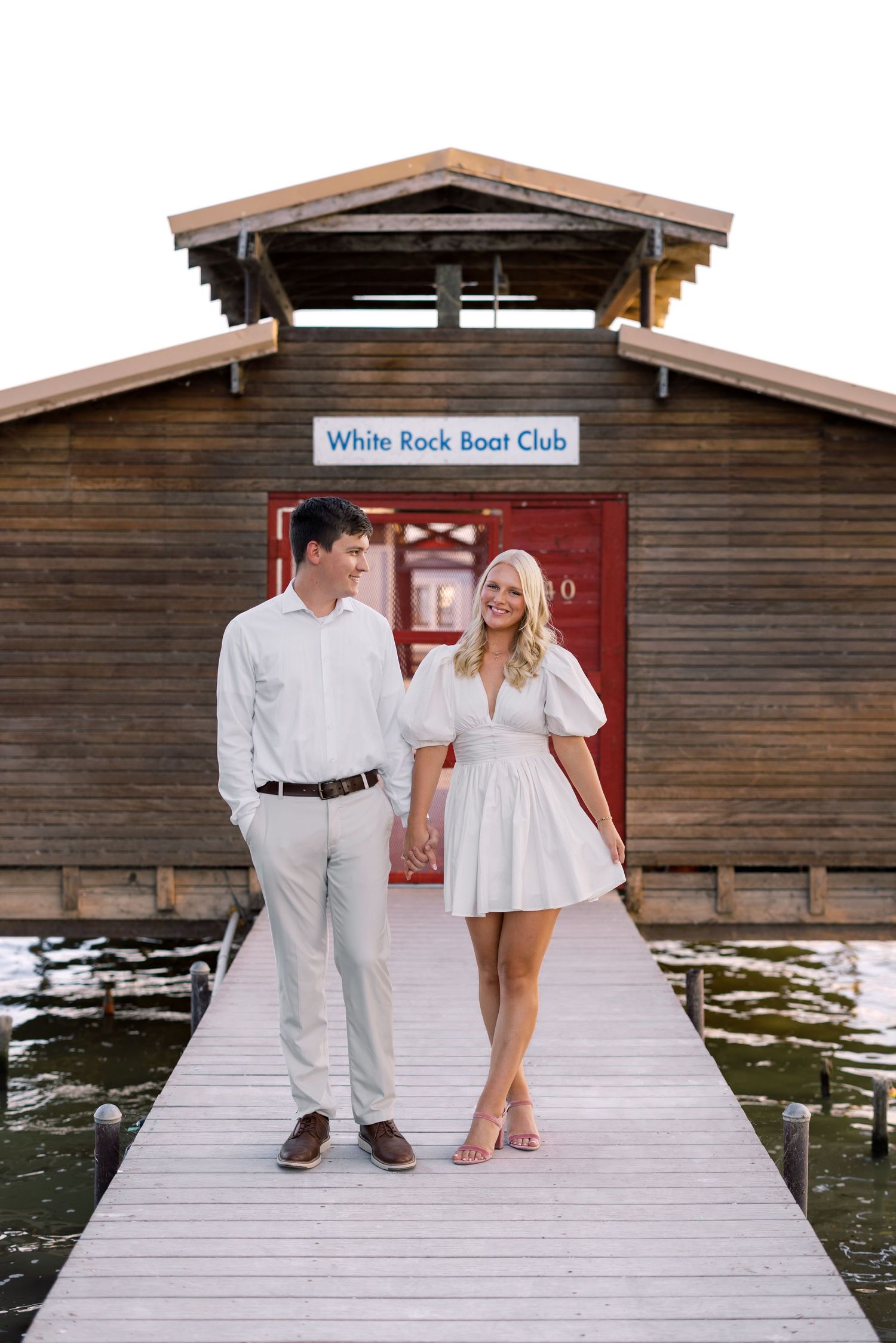 Couple holding hands on a dock in front of White Rock Boat Club, man in white shirt and woman in white mini dress.