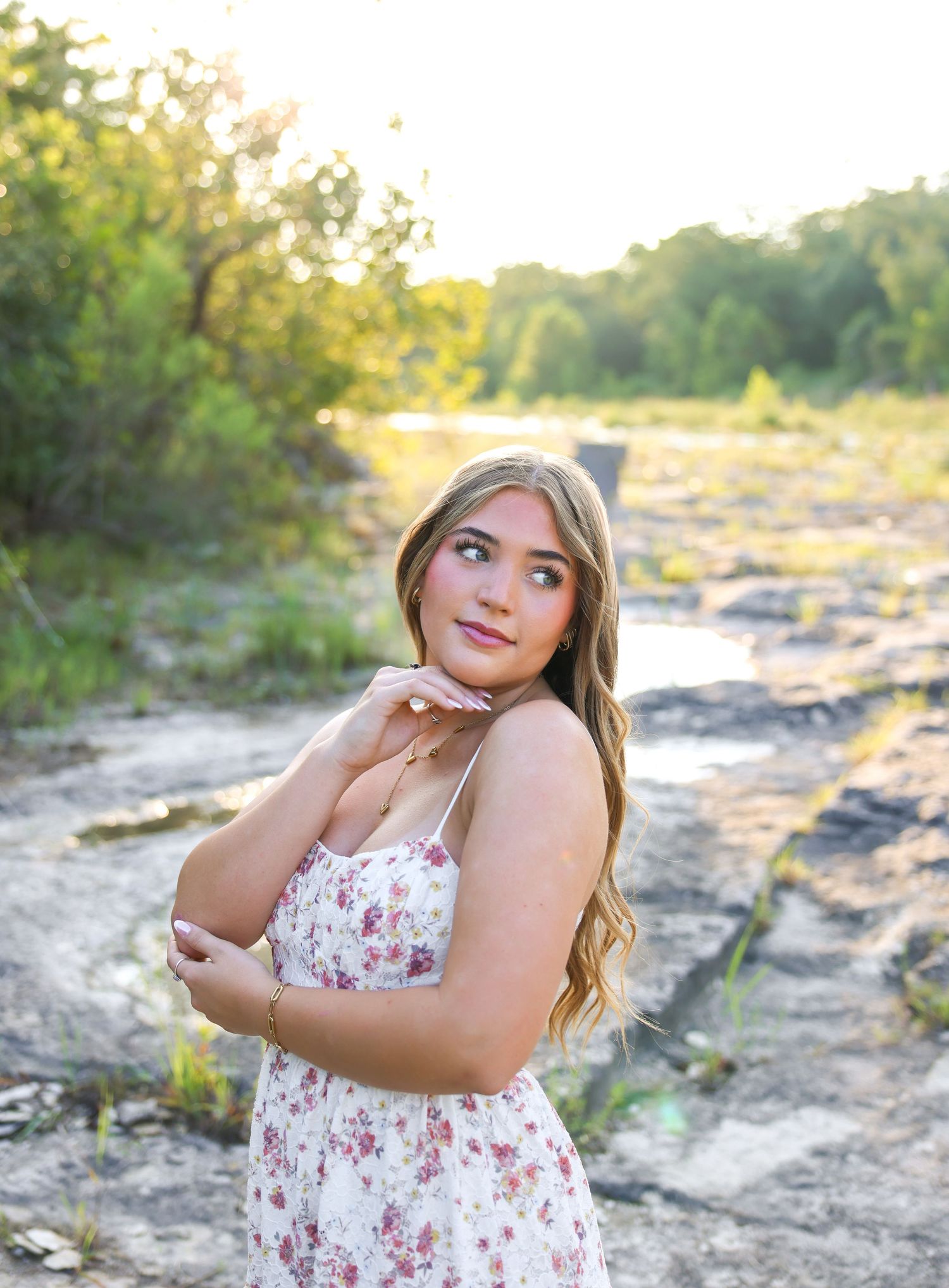 Young woman in floral sundress poses outdoors at golden hour near rocky creek bed surrounded by lush greenery.