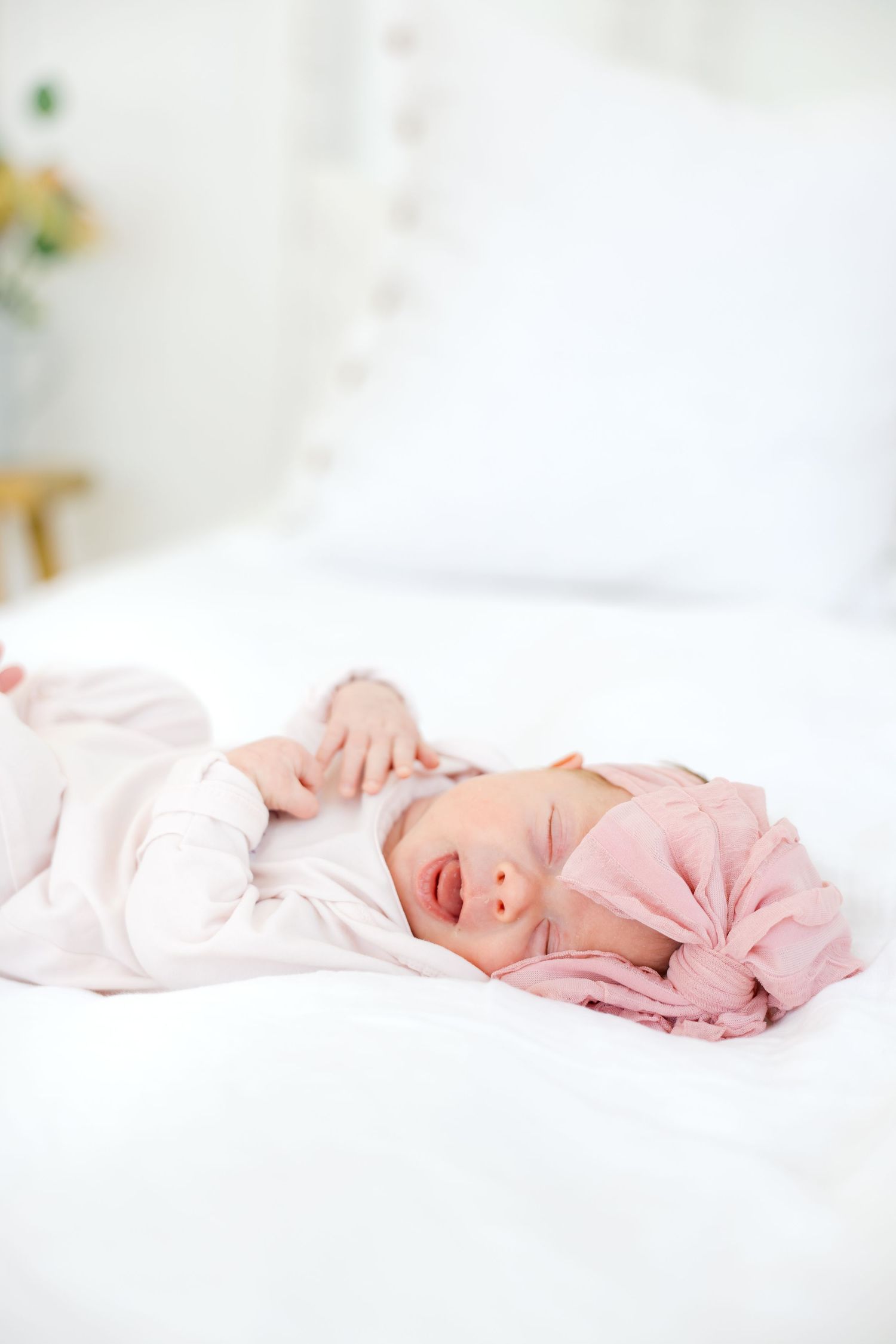 Newborn baby girl in pink outfit and bow headband lying on white bed with eyes slightly open.