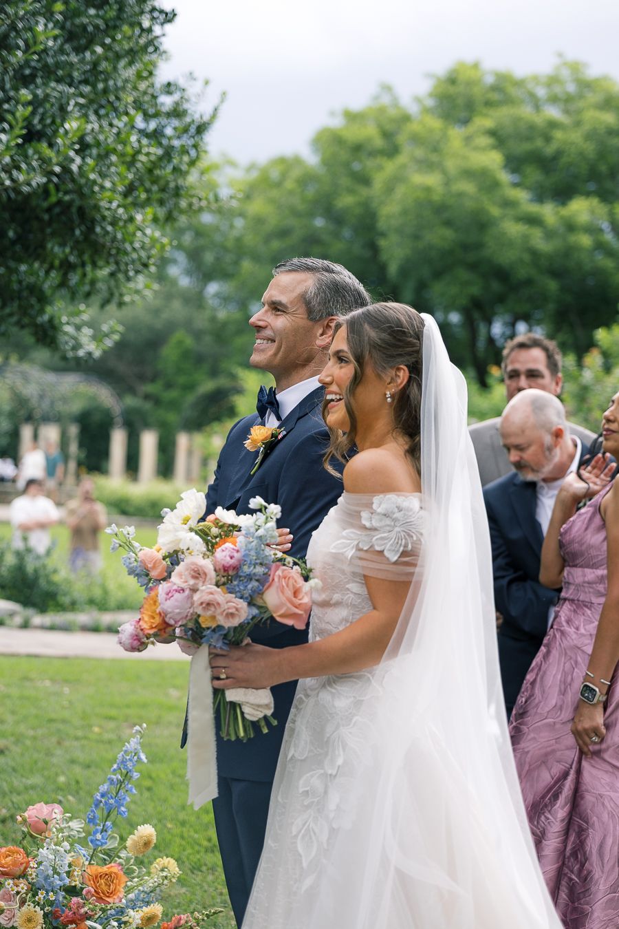 A radiant bride holding a colorful bouquet walks down the aisle with her escort at an outdoor wedding.