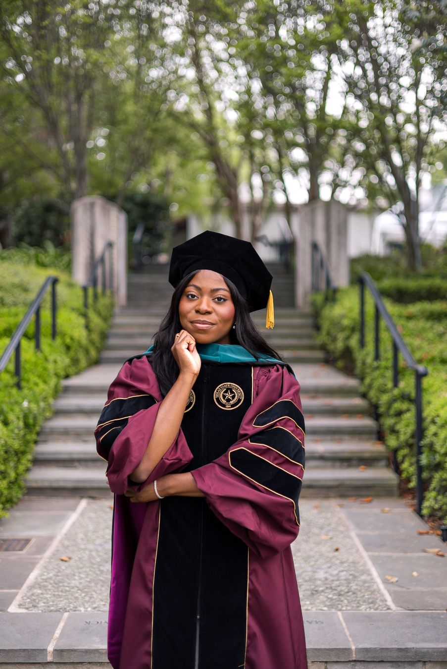 A young woman in maroon doctoral graduation regalia and black cap poses outdoors on a tree-lined pathway.