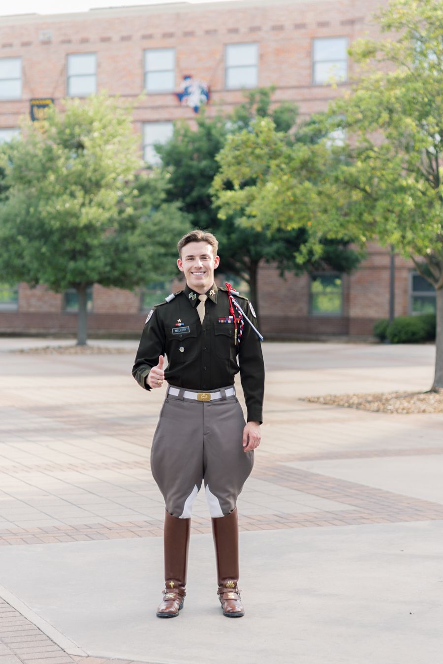 Young man in military dress uniform giving thumbs up outdoors near brick building on college campus.