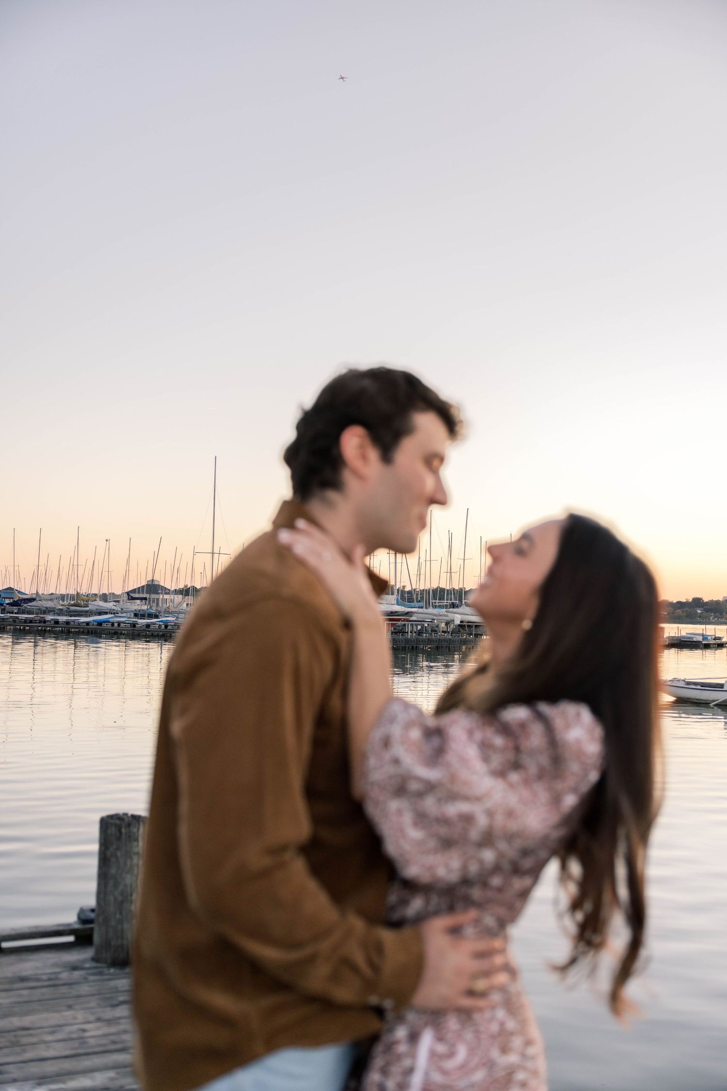 Romantic couple embracing on a wooden dock at sunset with a marina and sailboats in the background.