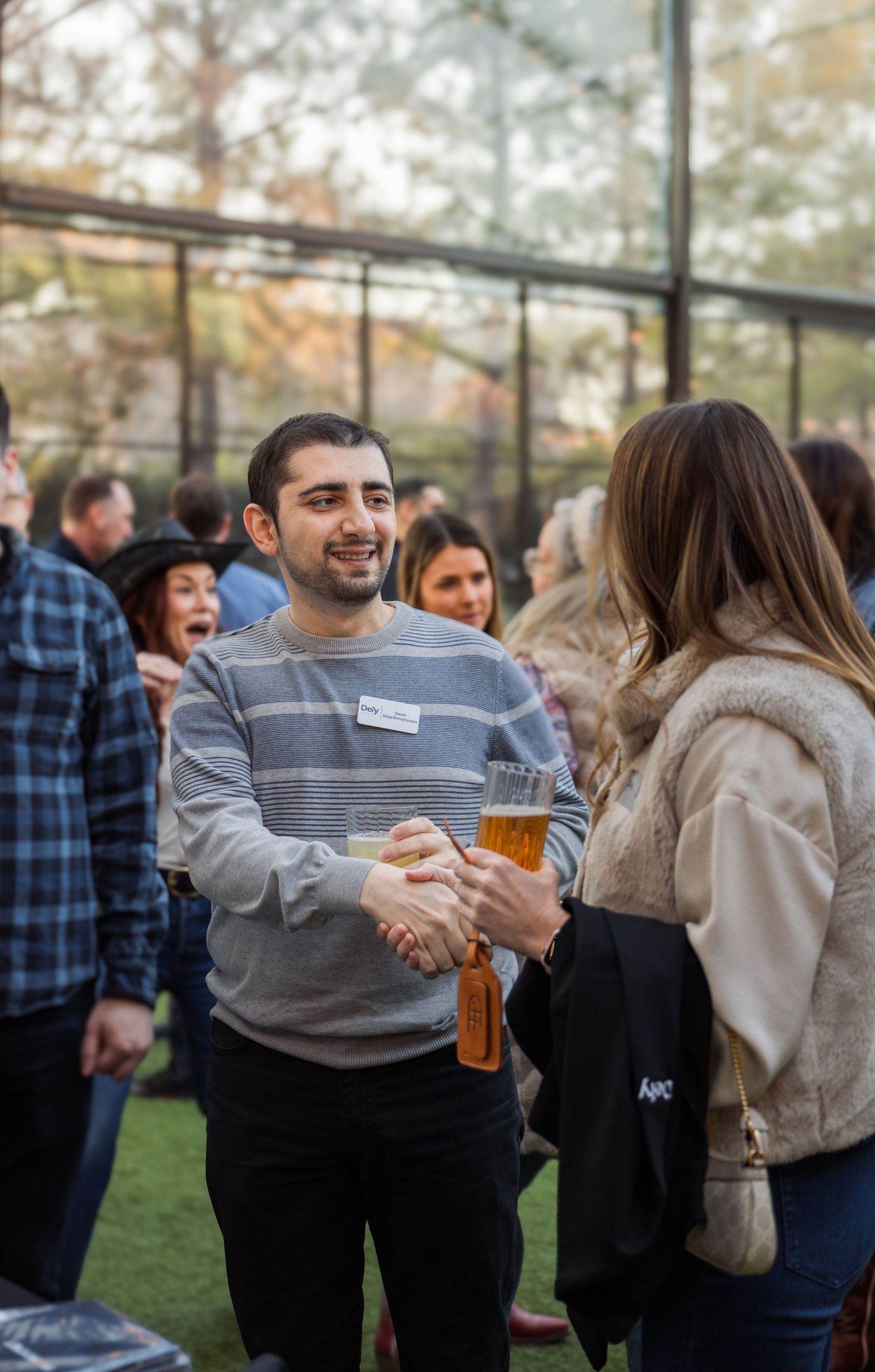 People socializing outdoors at a casual networking event, man in striped sweater holding beer and shaking hands.
