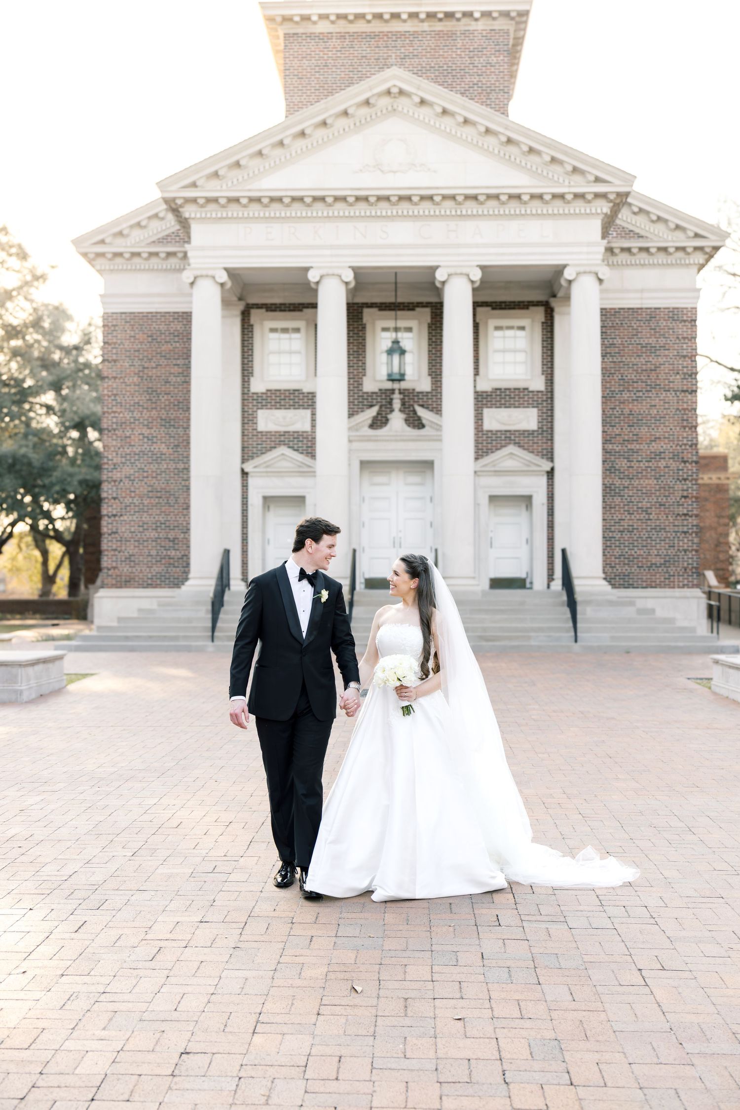 Bride and groom walk hand in hand in front of a grand historic brick building with white columns.