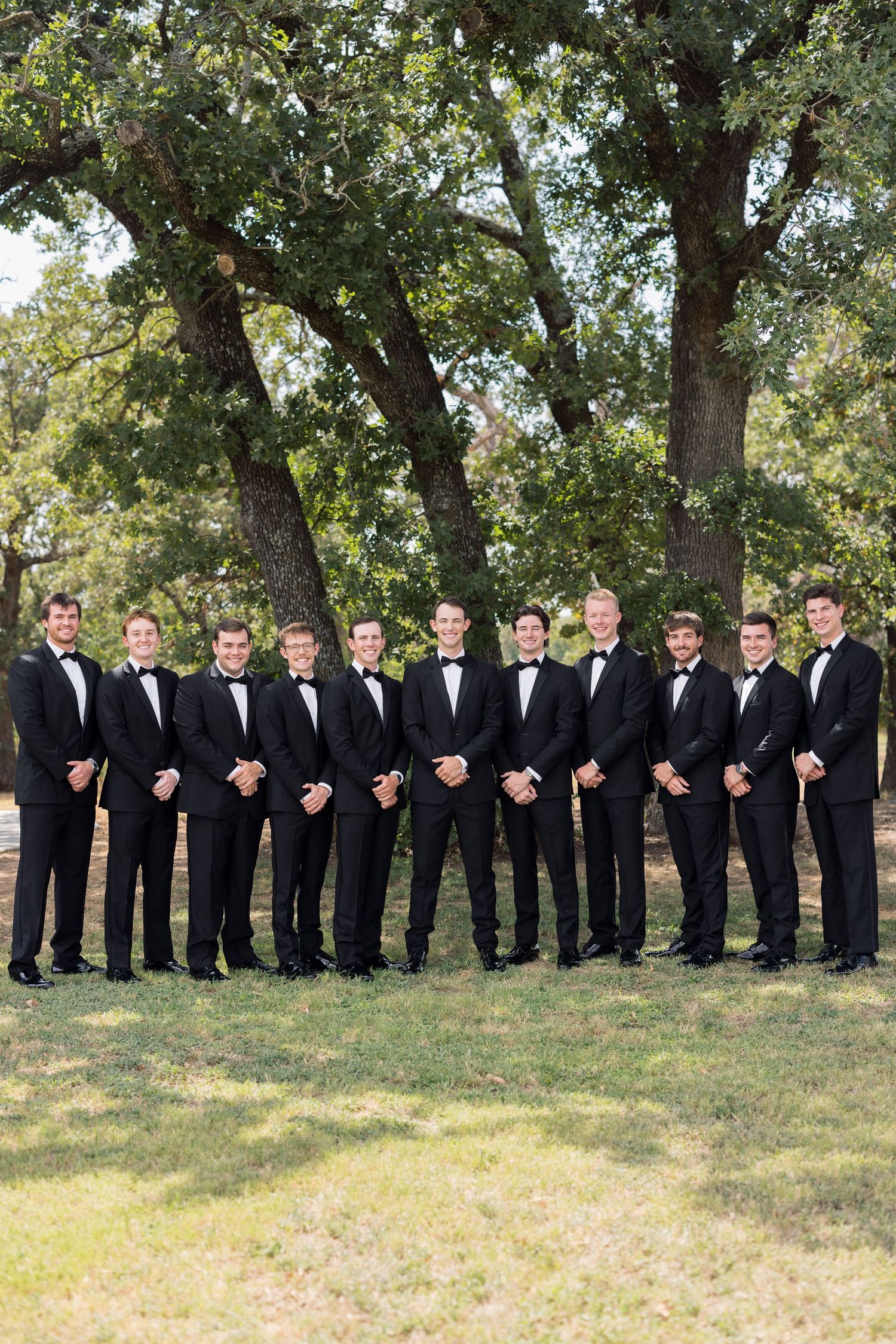 Groom and groomsmen in matching black tuxedos and bow ties posing outdoors under large oak trees on wedding day.