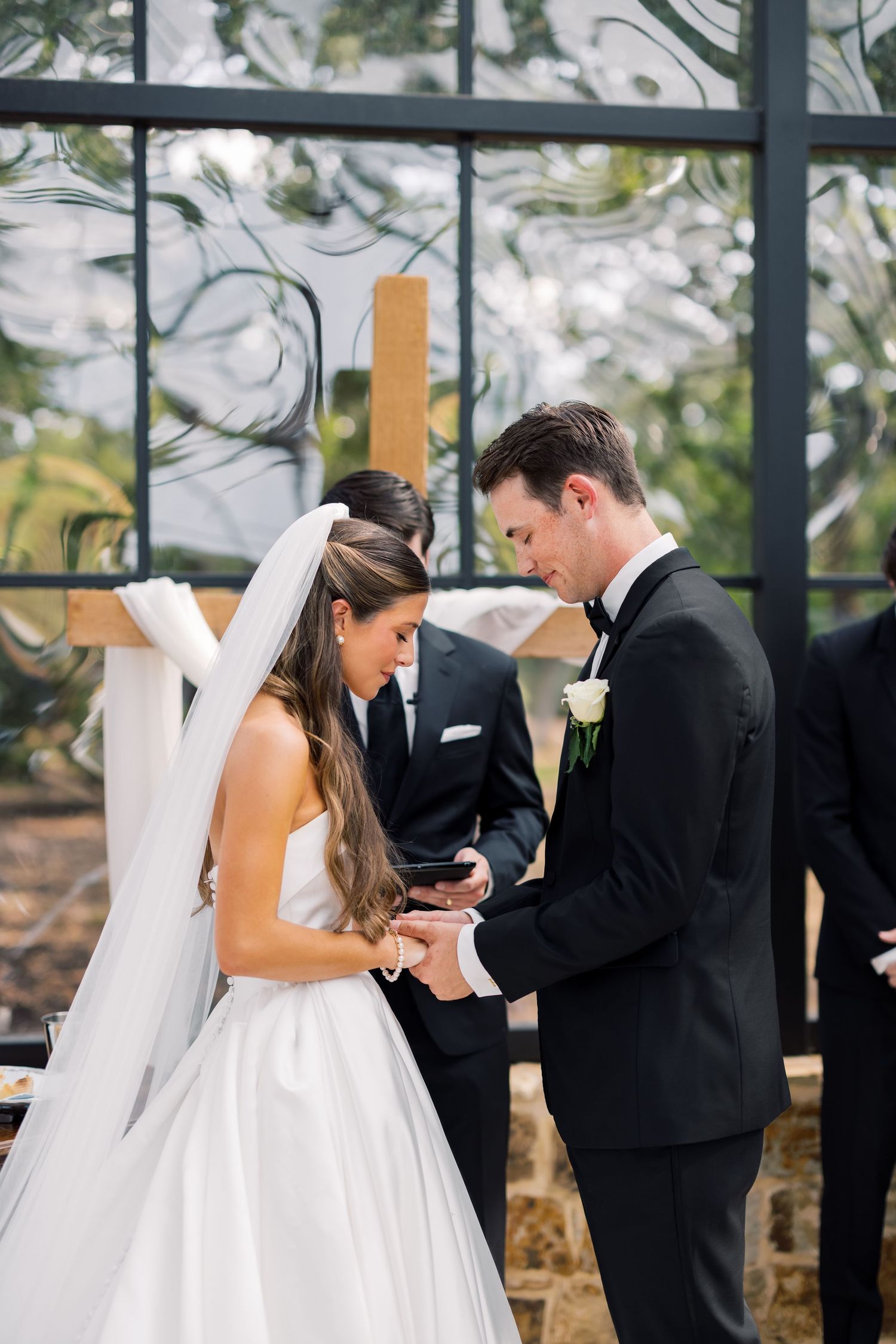 Bride and groom exchange rings at elegant wedding ceremony before large windows with wooden cross.