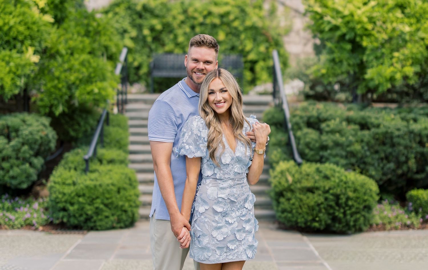 Happy couple posing outdoors in coordinating blue outfits, smiling in a lush green garden setting.