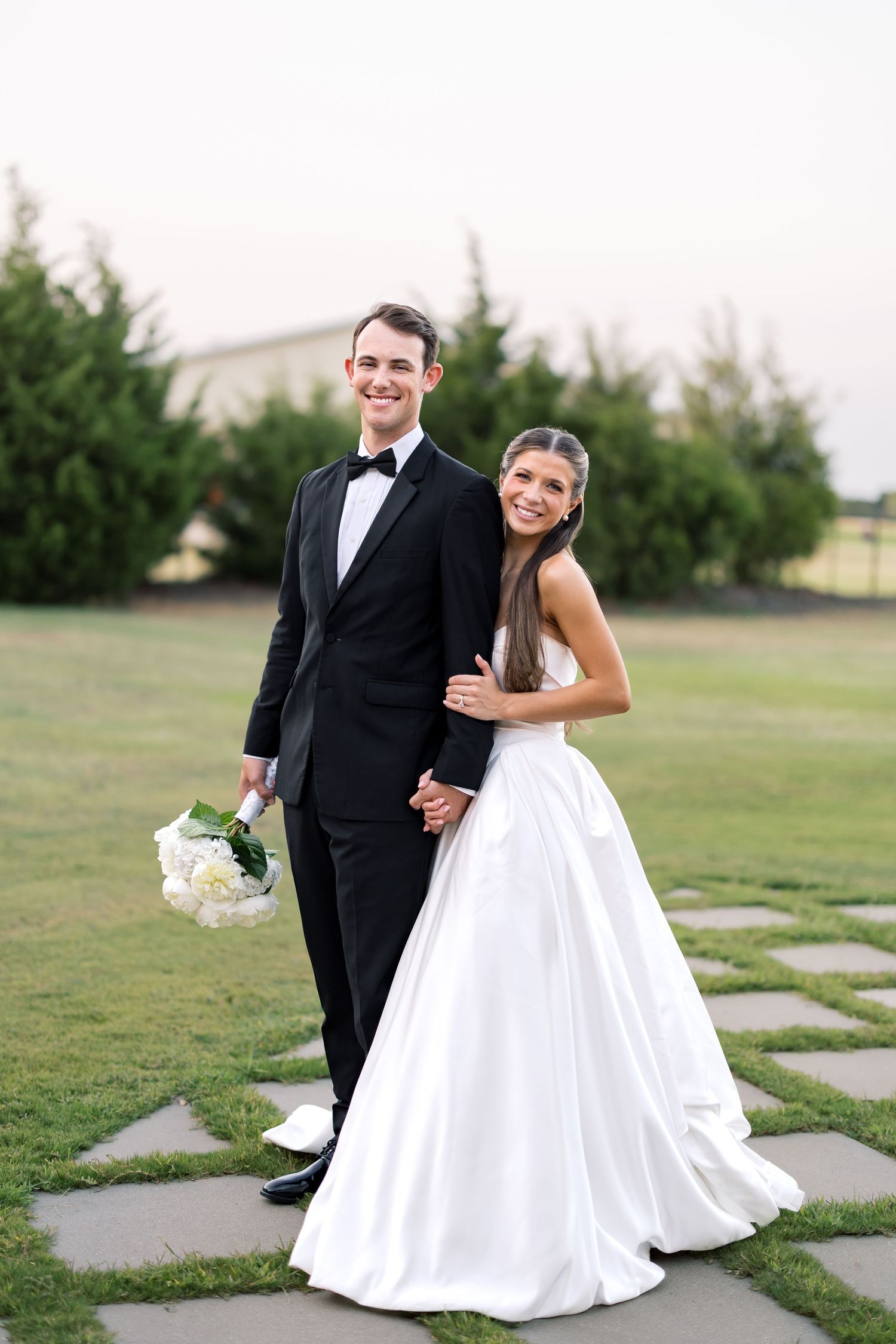 Smiling bride in white ballgown and groom in black tuxedo holding white bouquet pose on stone path outdoors.