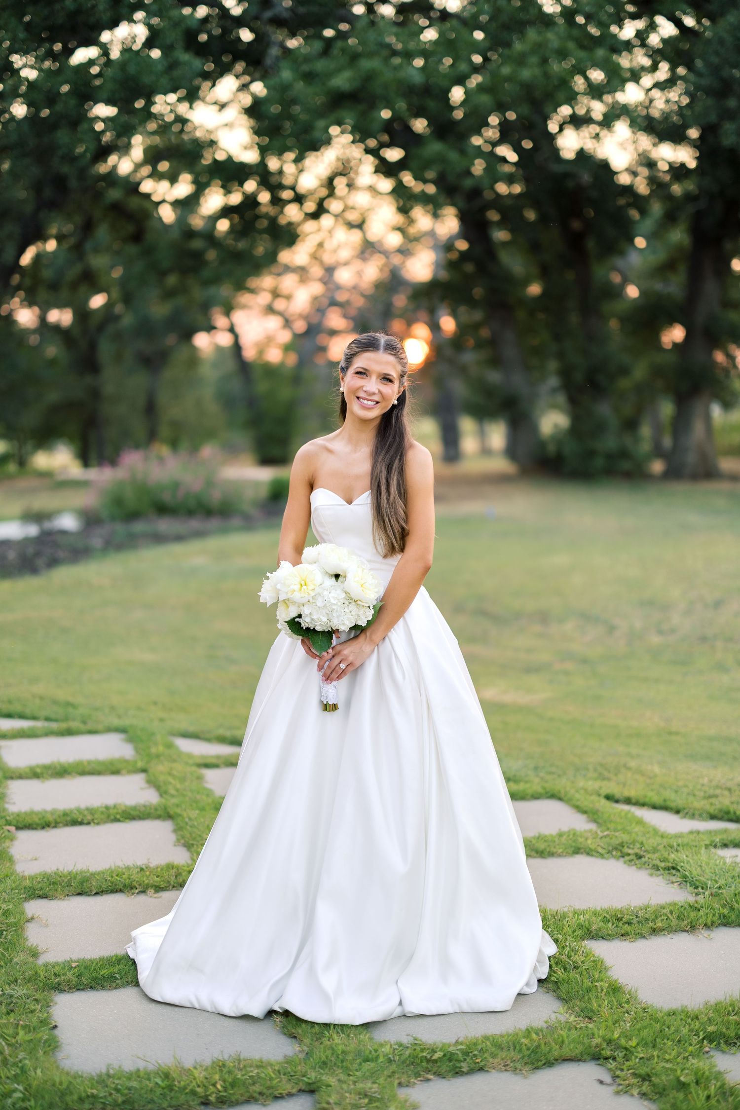 Smiling bride in strapless white ball gown holds white bouquet on stone path in lush garden at golden hour.