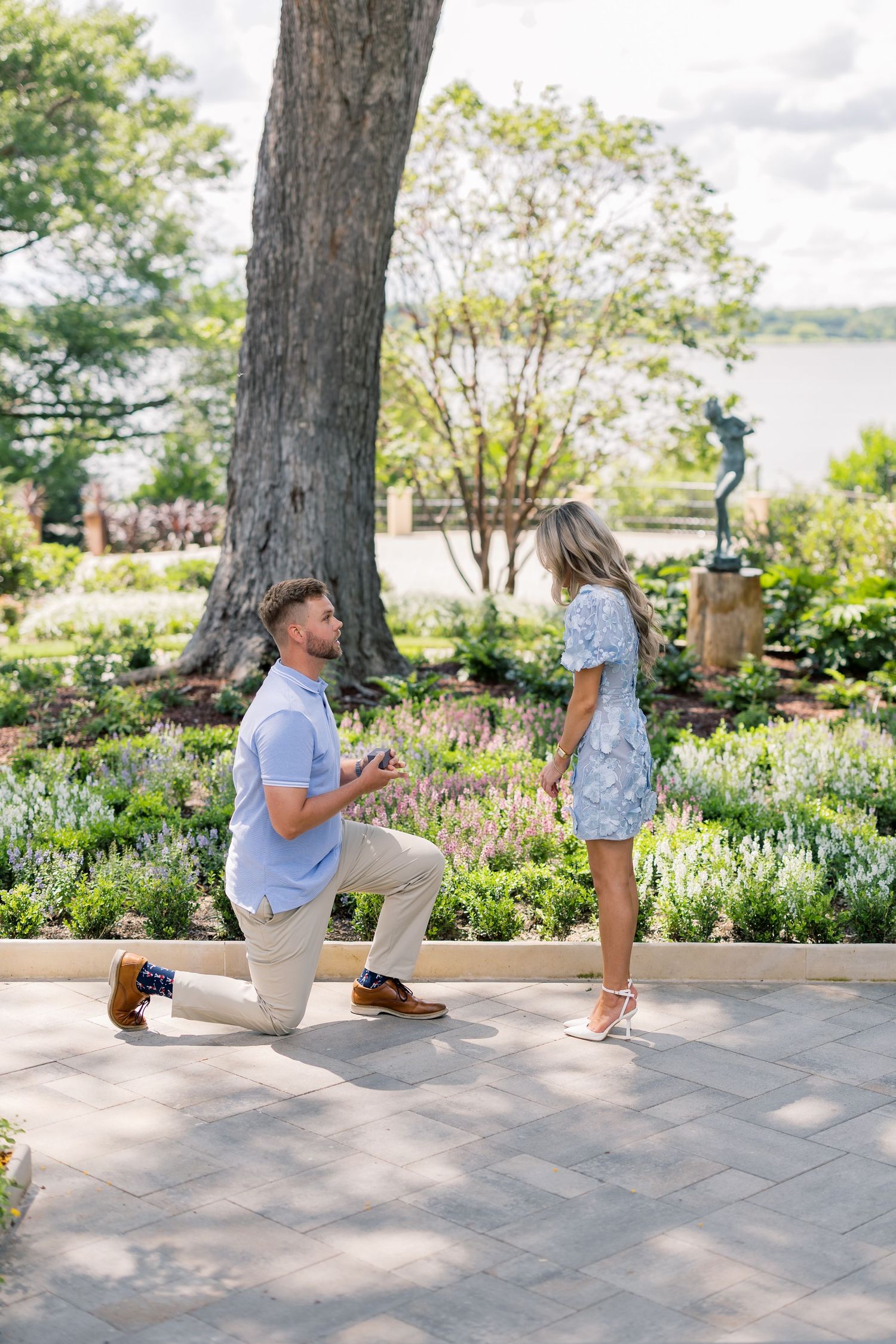 Man proposes on one knee to woman in floral dress in a scenic garden with lush greenery and water in background.