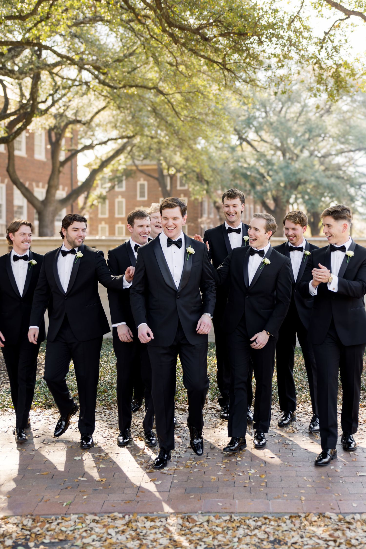 Smiling groom walks with groomsmen in matching black tuxedos on a tree-lined brick path.