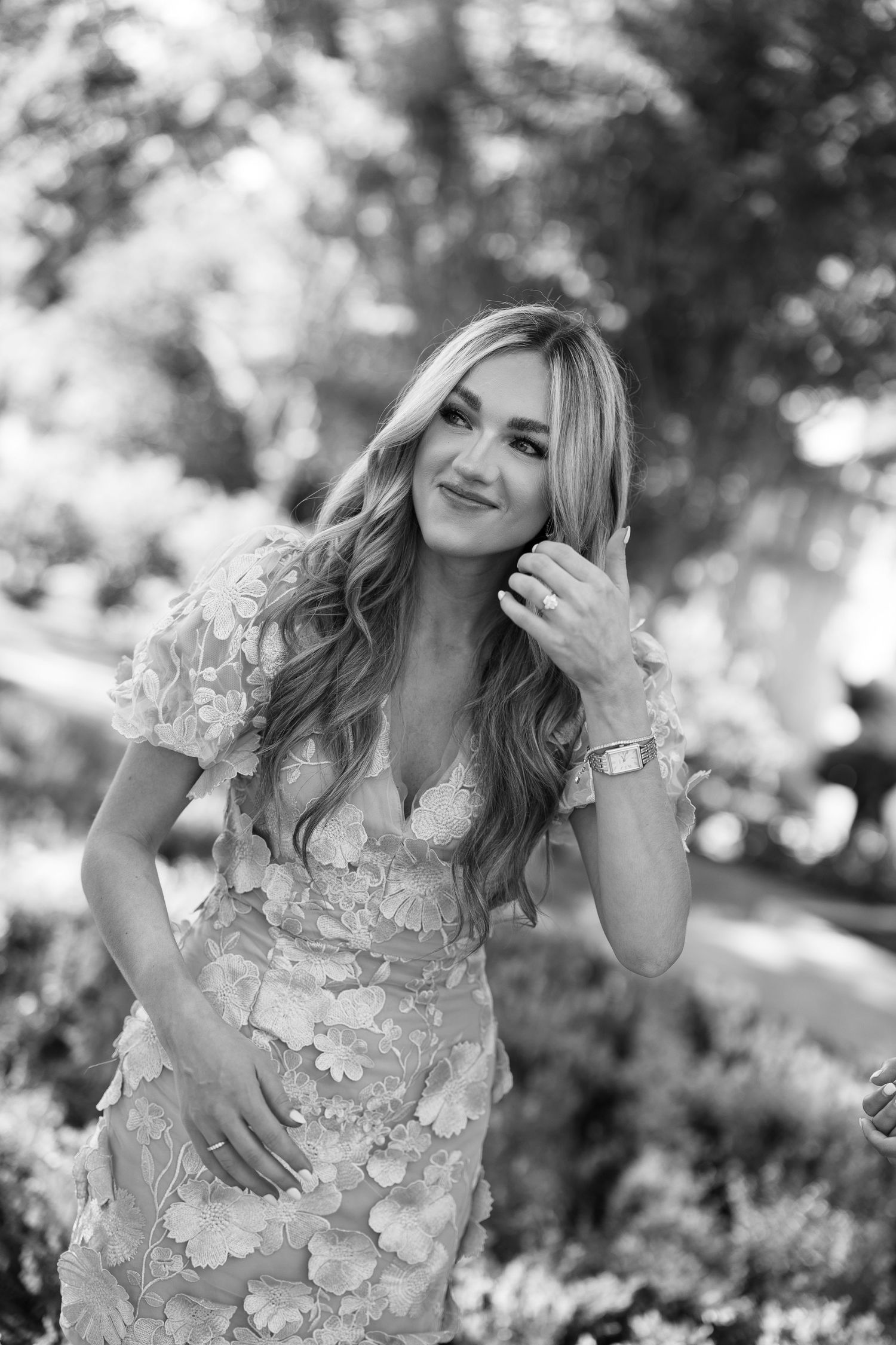 Black and white portrait of a smiling blonde woman in a floral dress posing outdoors in a garden setting.