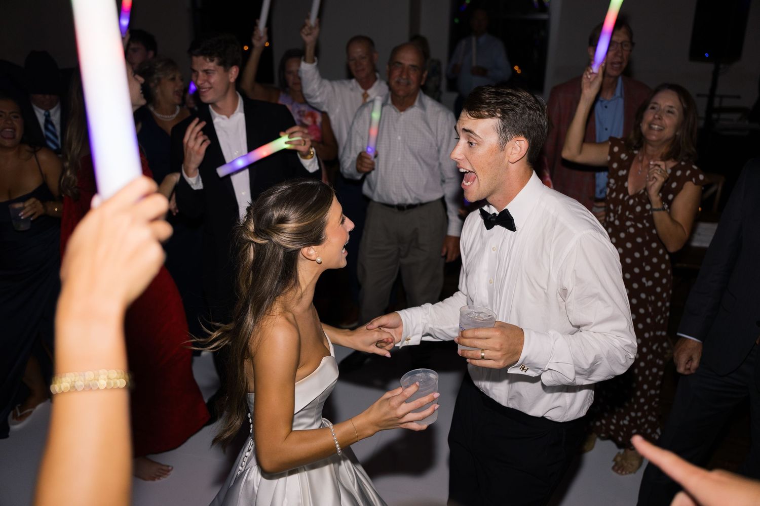 Bride and groom dancing joyfully with guests holding colorful glow sticks at a lively wedding reception.