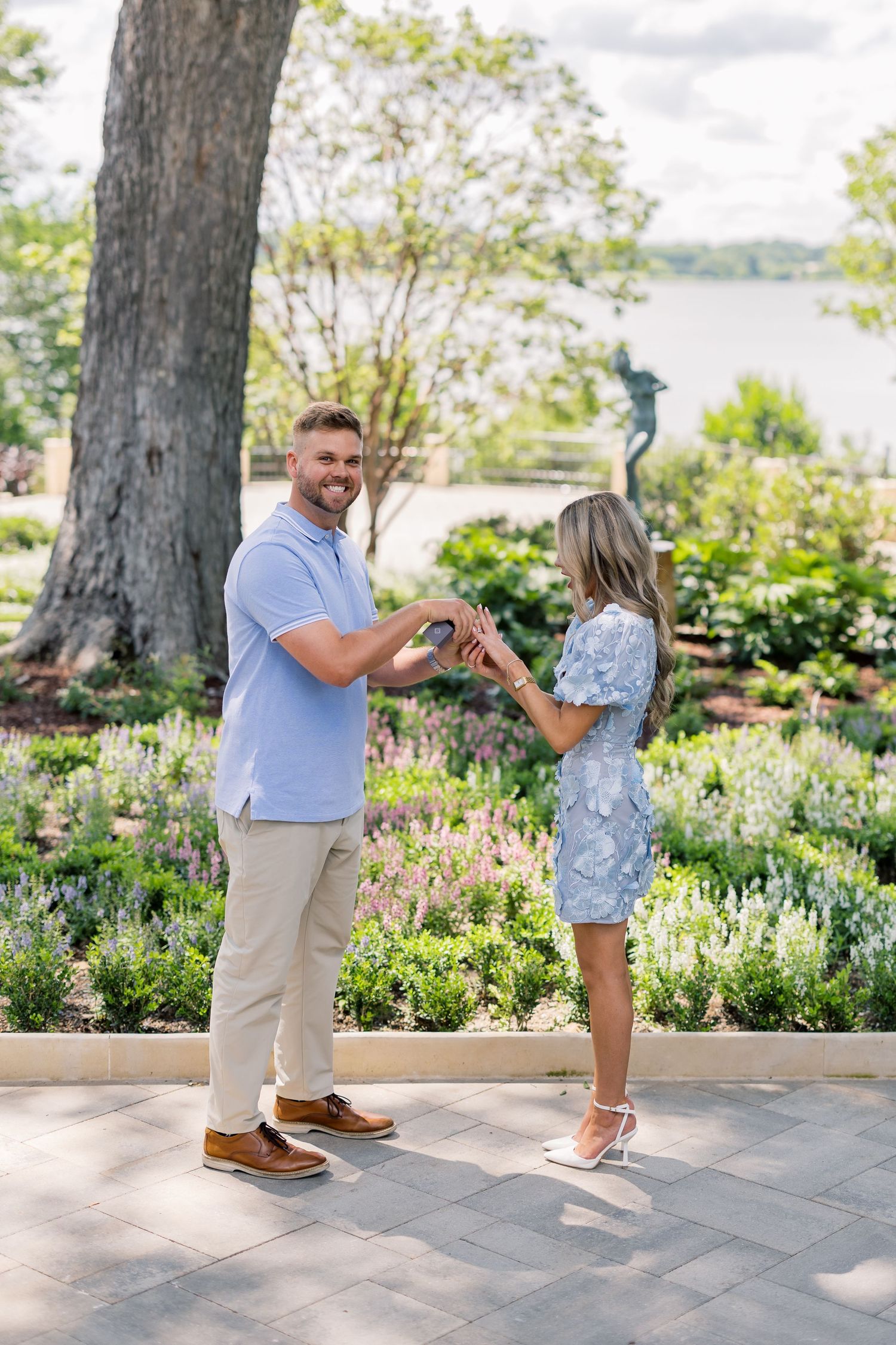 A couple holds hands in a lush garden near a lake, the man in a blue polo and the woman in a floral dress.