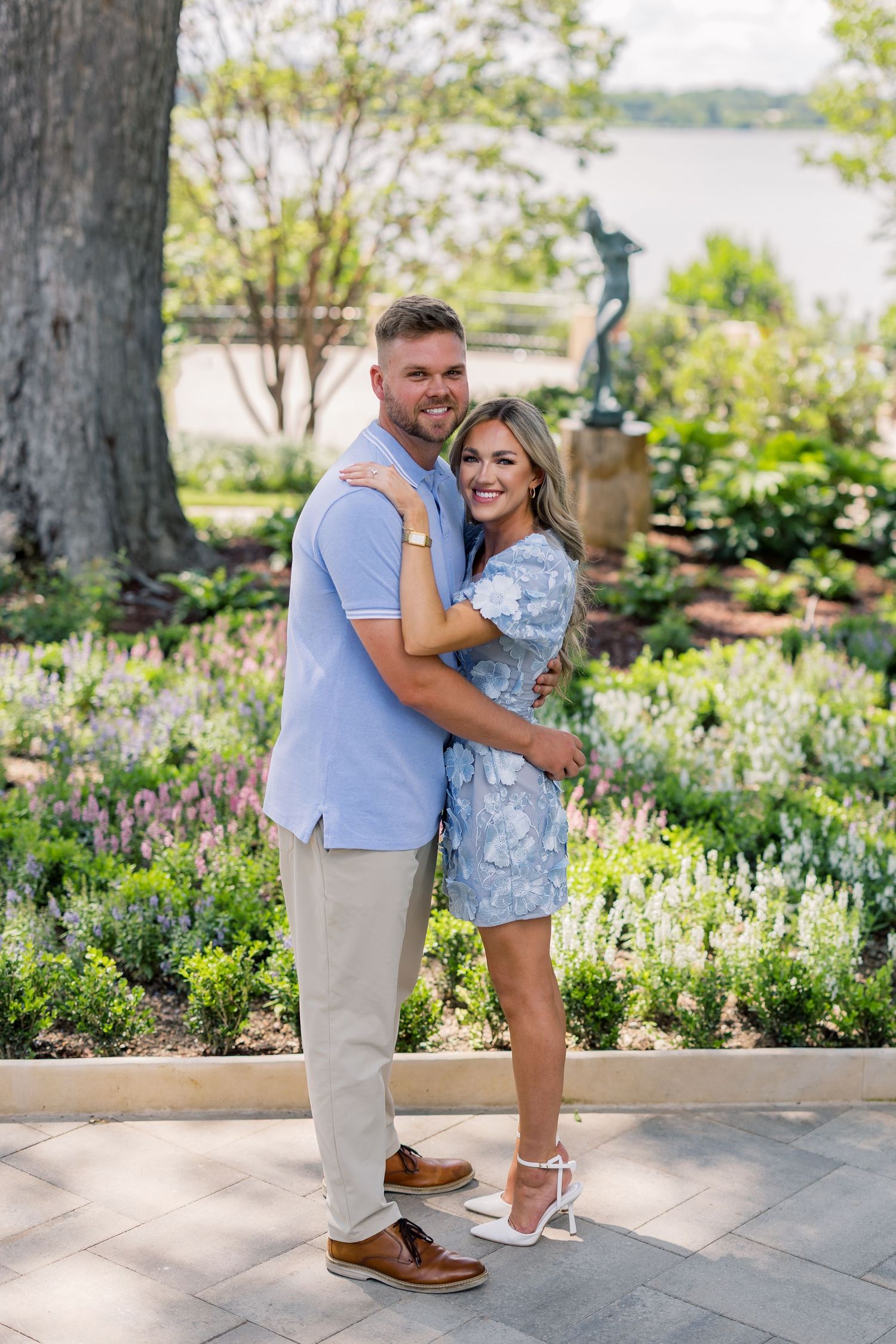 A couple embraces and smiles in a lush garden with colorful flowers and a water view behind them.