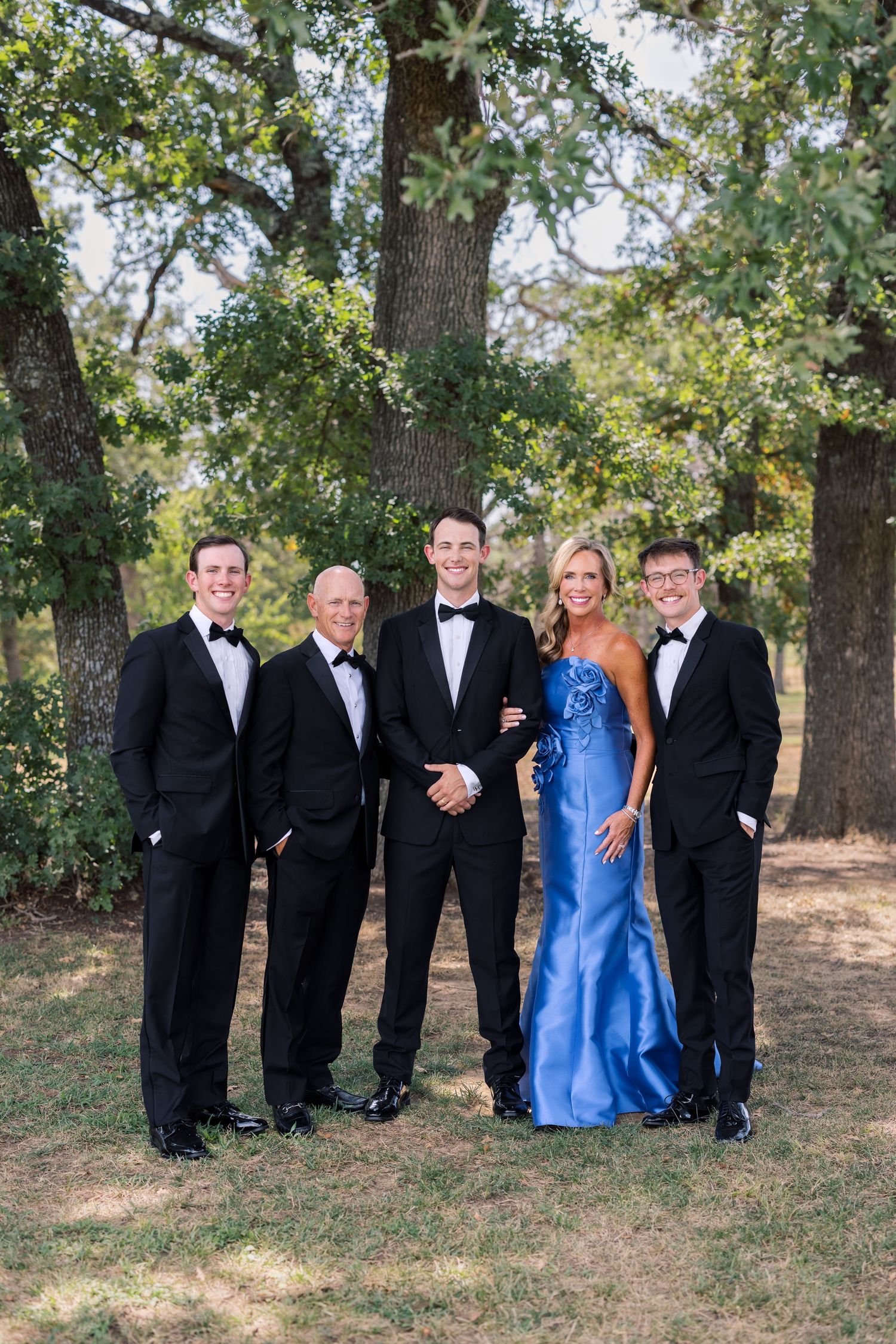 Group of five posing outdoors, four in black tuxedos and one woman in a blue gown, smiling among trees.