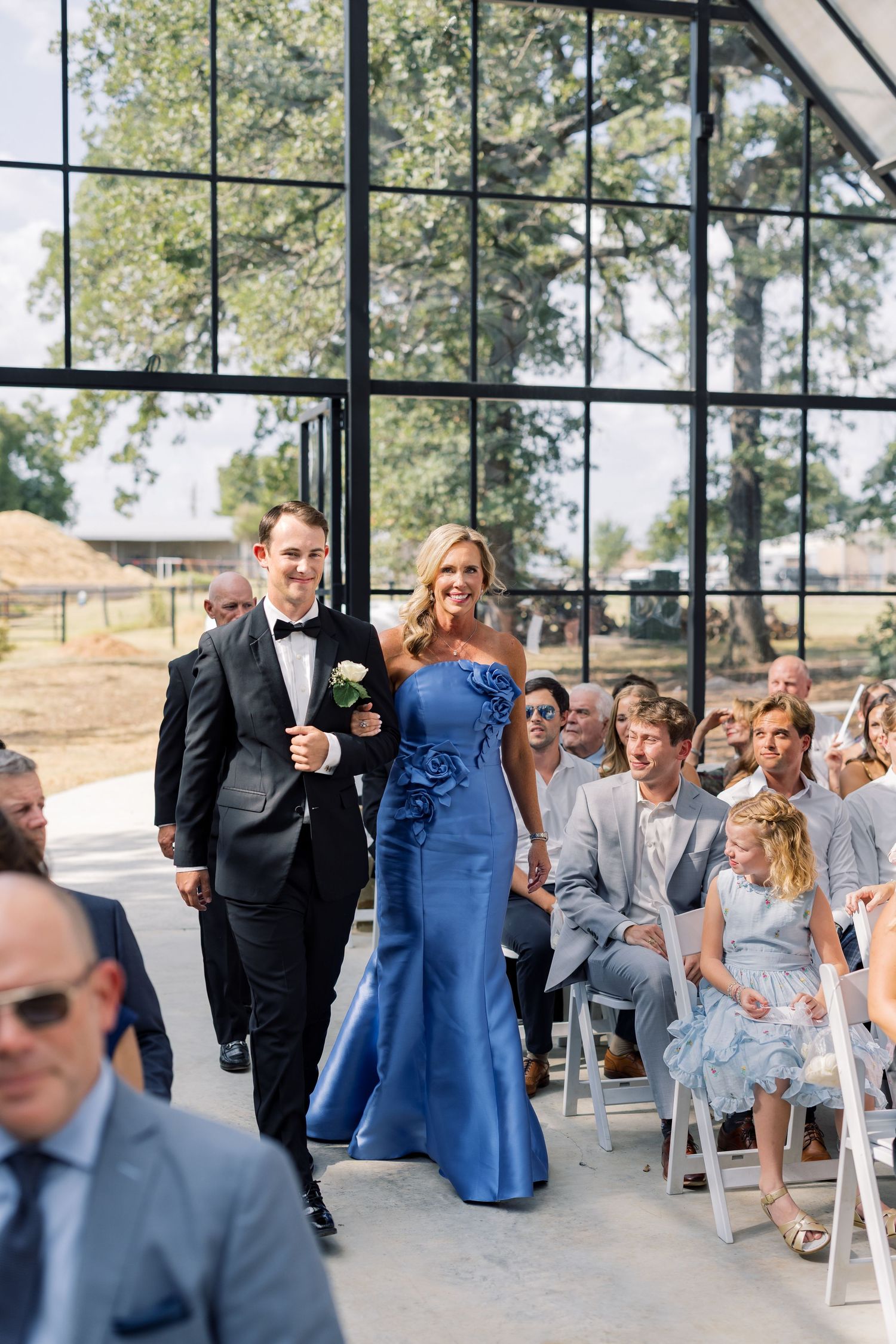 A groomsman in a black tuxedo escorts a woman in a blue strapless gown down the aisle at an elegant wedding ceremony.