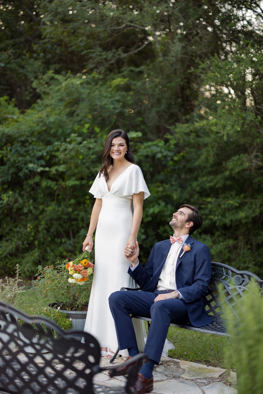 Bride in white flutter-sleeve gown holds bouquet standing beside groom in navy suit with pink bow tie on garden bench.