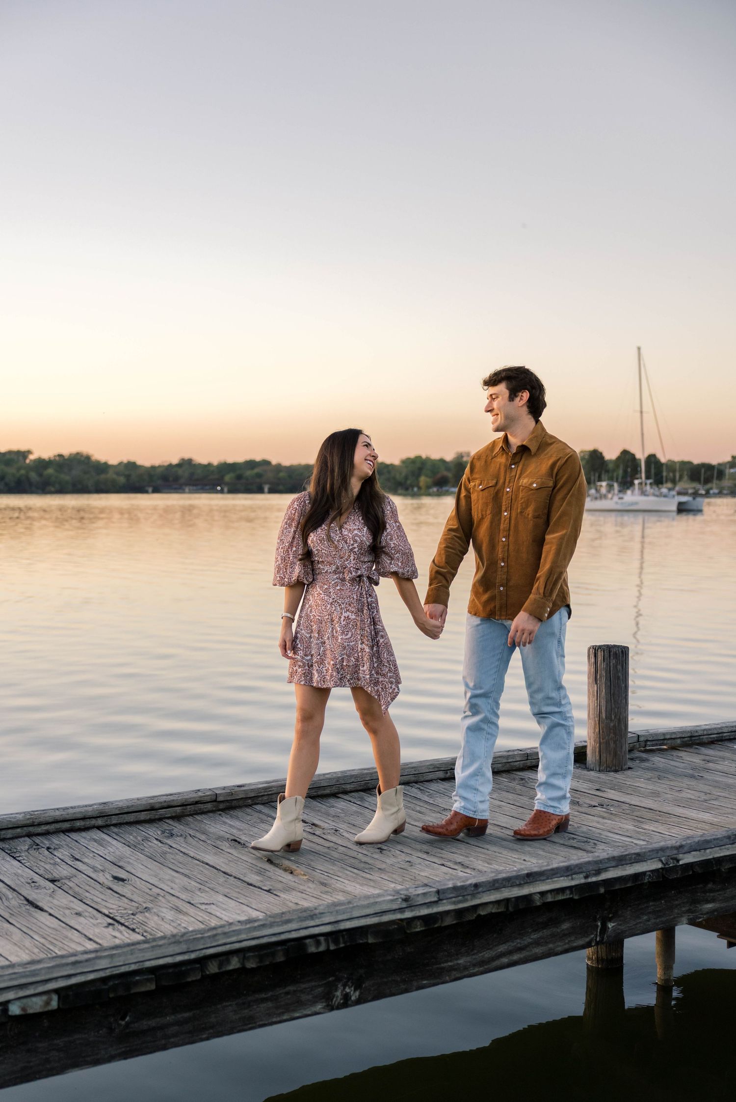 Couple holding hands on a wooden dock at sunset by a calm lake, woman in floral dress and man in brown shirt and jeans.
