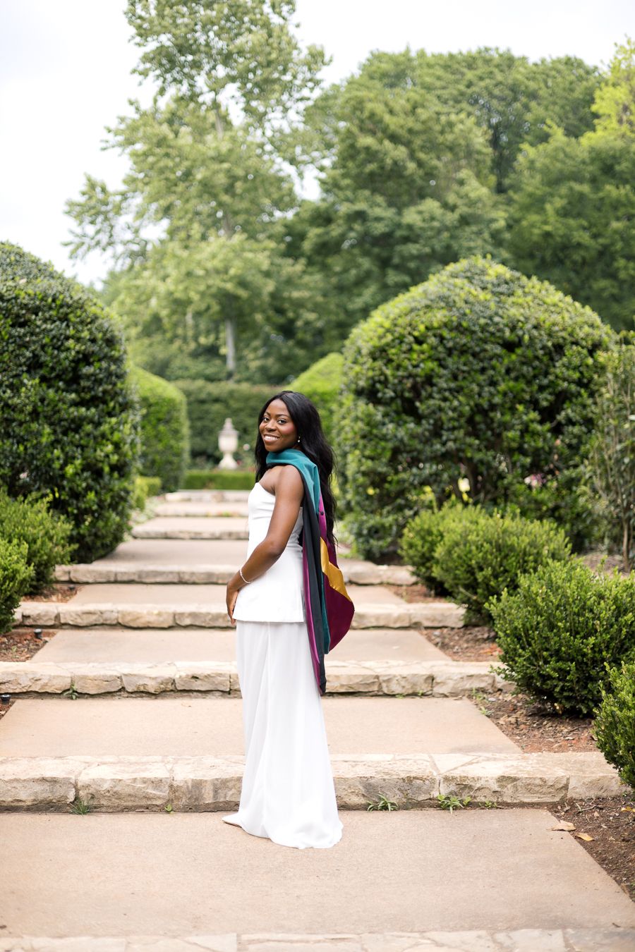 A woman in a white outfit and colorful scarf poses on stone steps in a lush green garden.