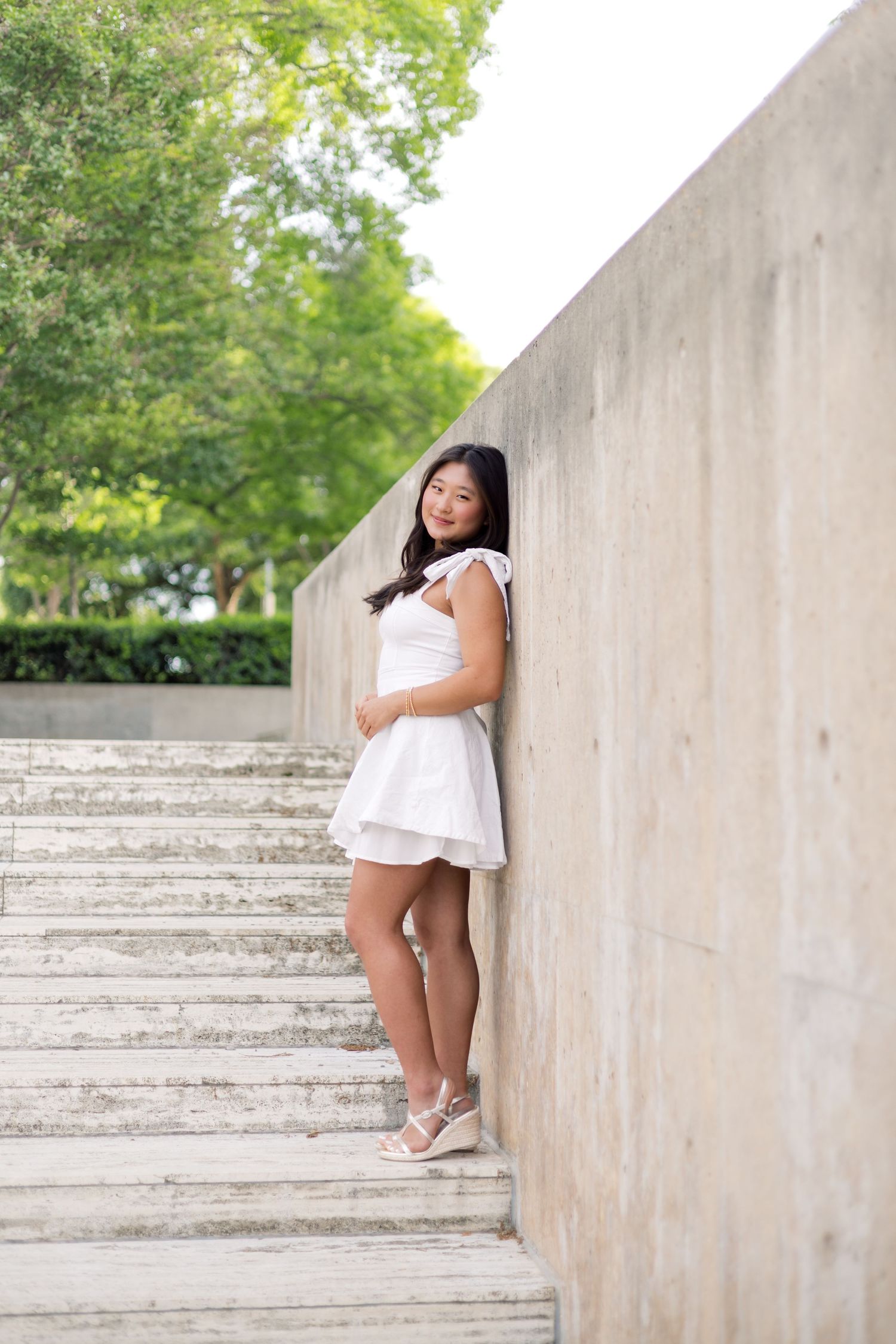 Young Asian woman in white dress posing gracefully on outdoor concrete steps beside a wall.