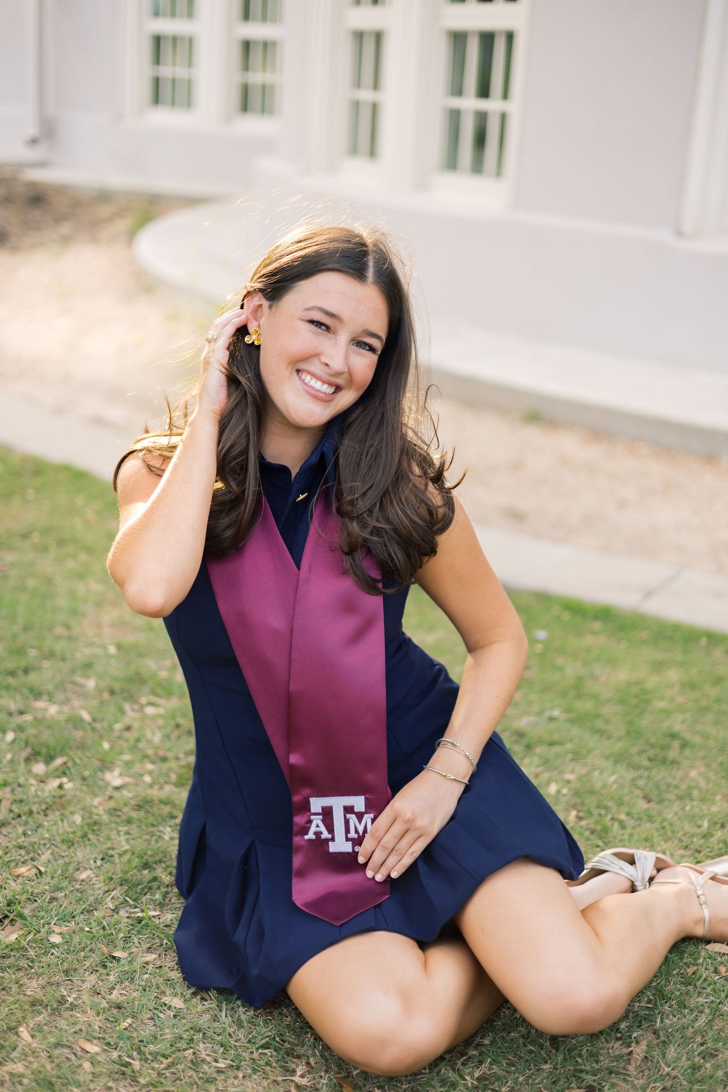 Smiling Texas A&M graduate sits on grass in navy dress with maroon ATM stole, fixing her hair.