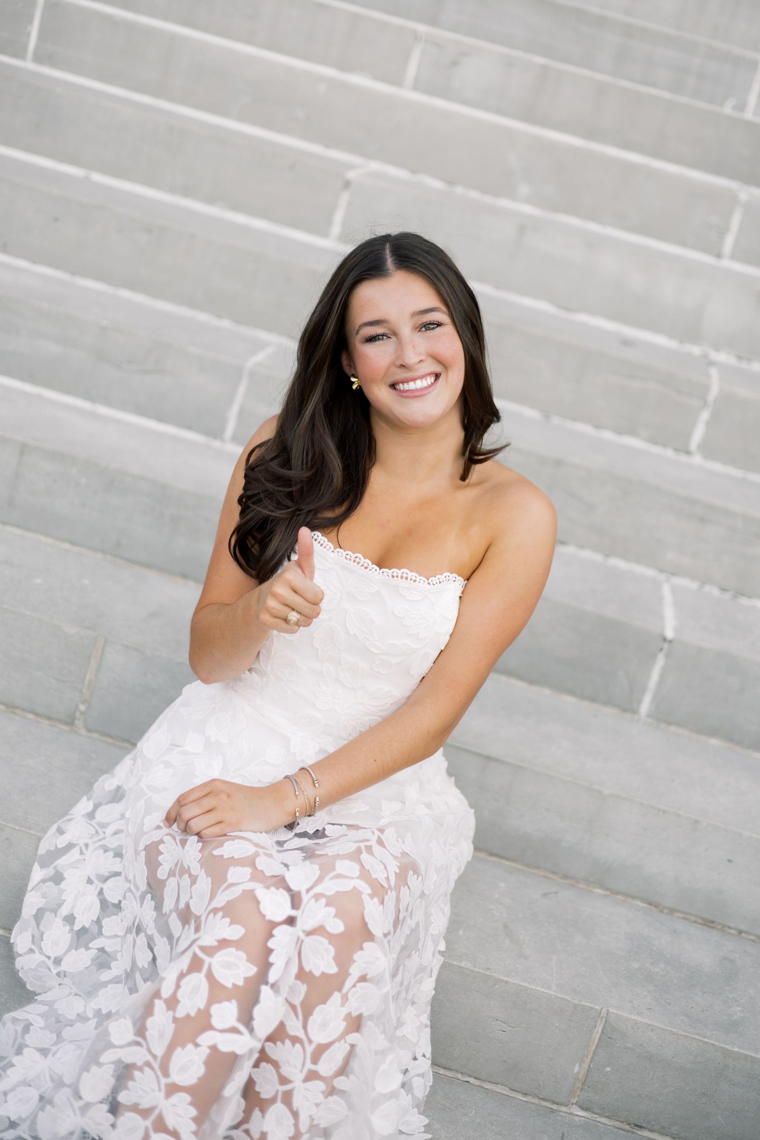 Happy brunette in strapless white floral lace dress giving thumbs up on marble steps.