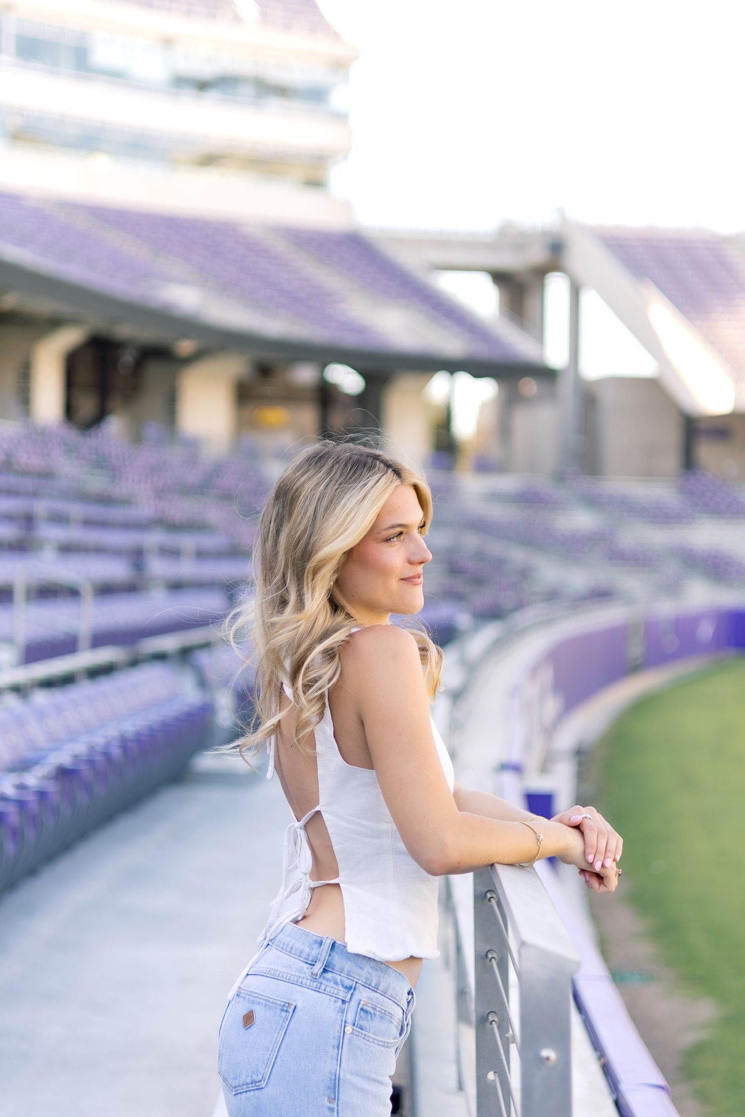 Blonde woman in white top and jeans gazing back while leaning on stadium railing.