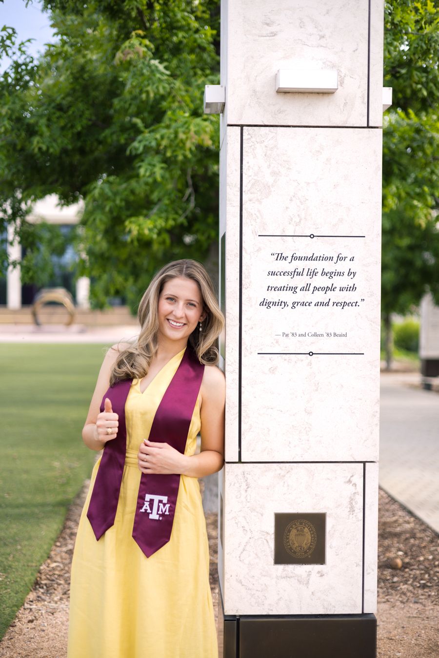 Texas A&M graduate in yellow dress and maroon stole gives thumbs up beside campus pillar with inspirational quote.