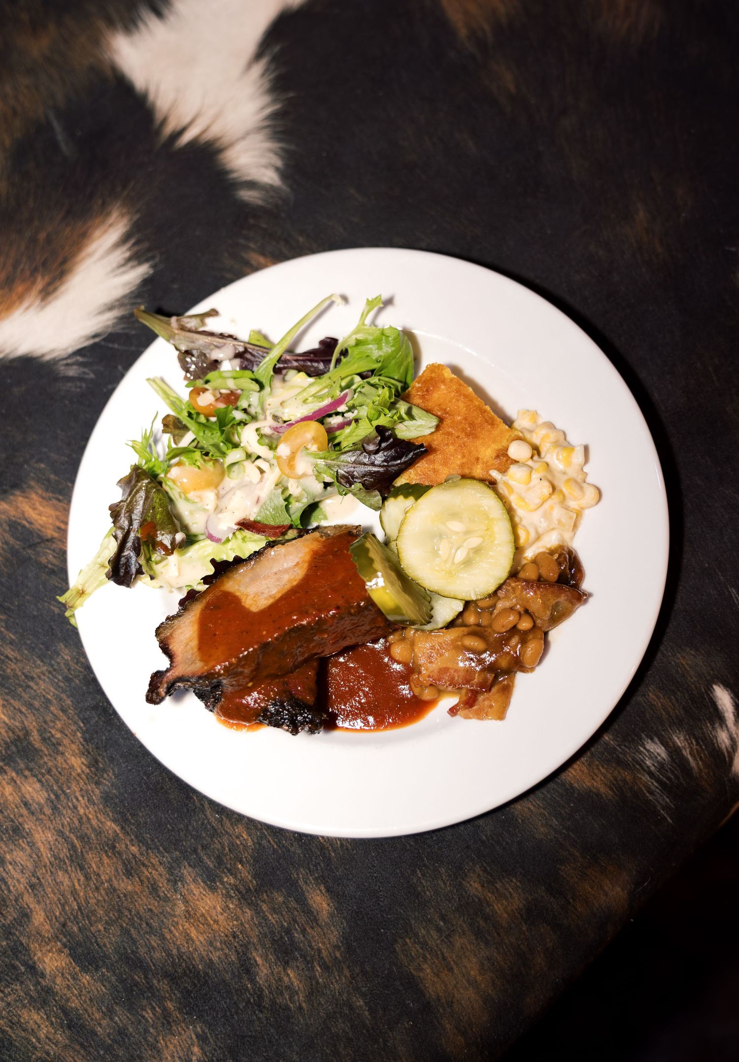BBQ plate with smoked meat, baked beans, cucumber slices, cornbread, and fresh mixed greens salad on white plate.