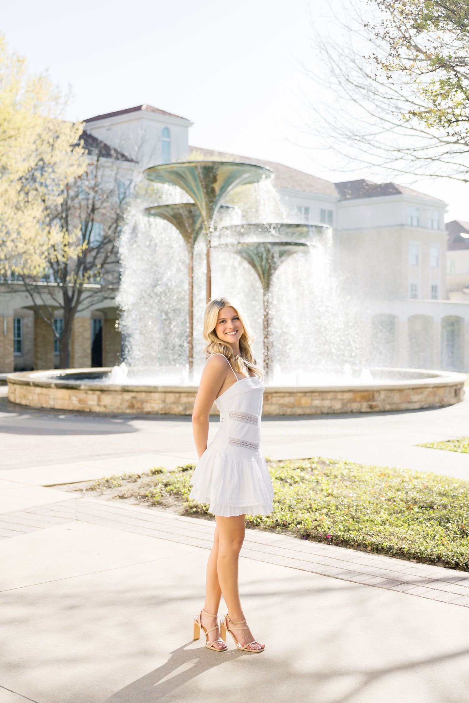 Smiling blonde woman in white dress poses in front of decorative fountain on sunny college campus.