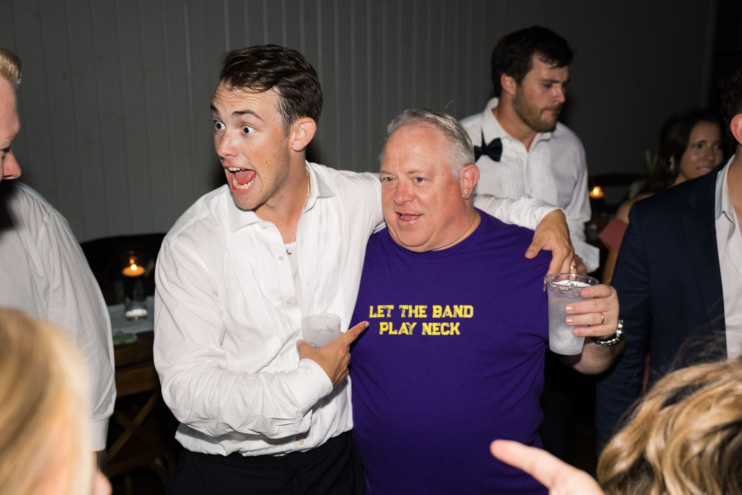 Two men pose and laugh at a party, one wearing a purple Let The Band Play Neck t-shirt while holding a drink.