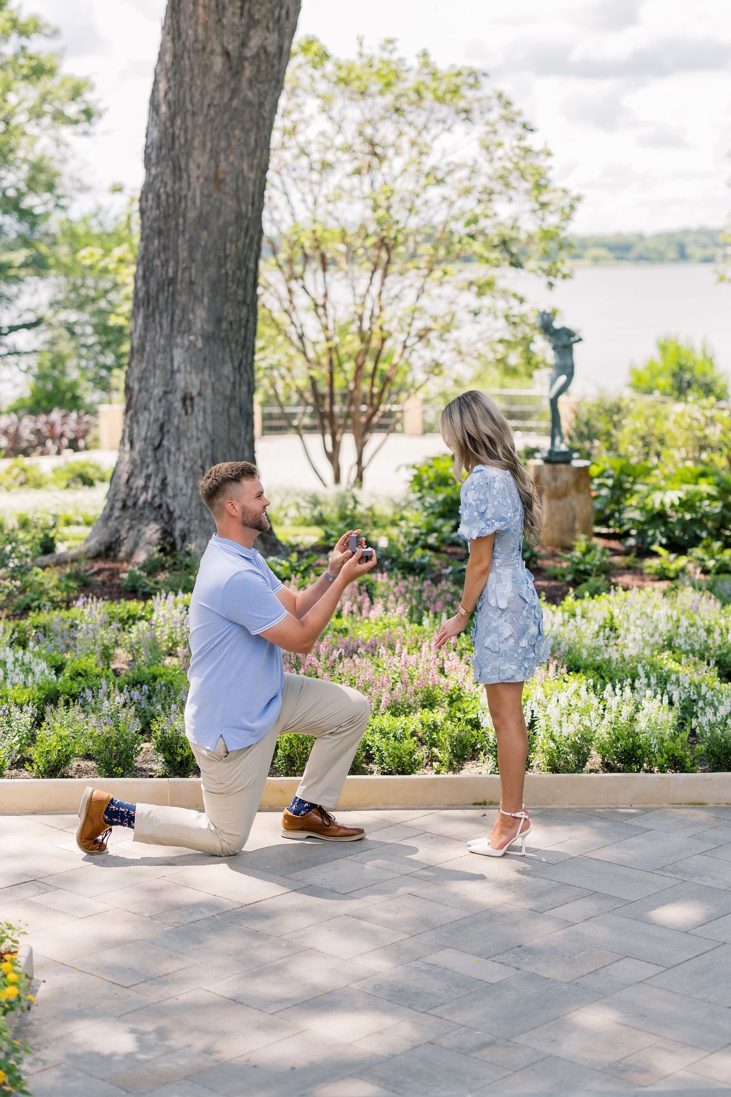 Man proposes on one knee to woman in floral dress in a lush garden near a waterfront on a sunny day.