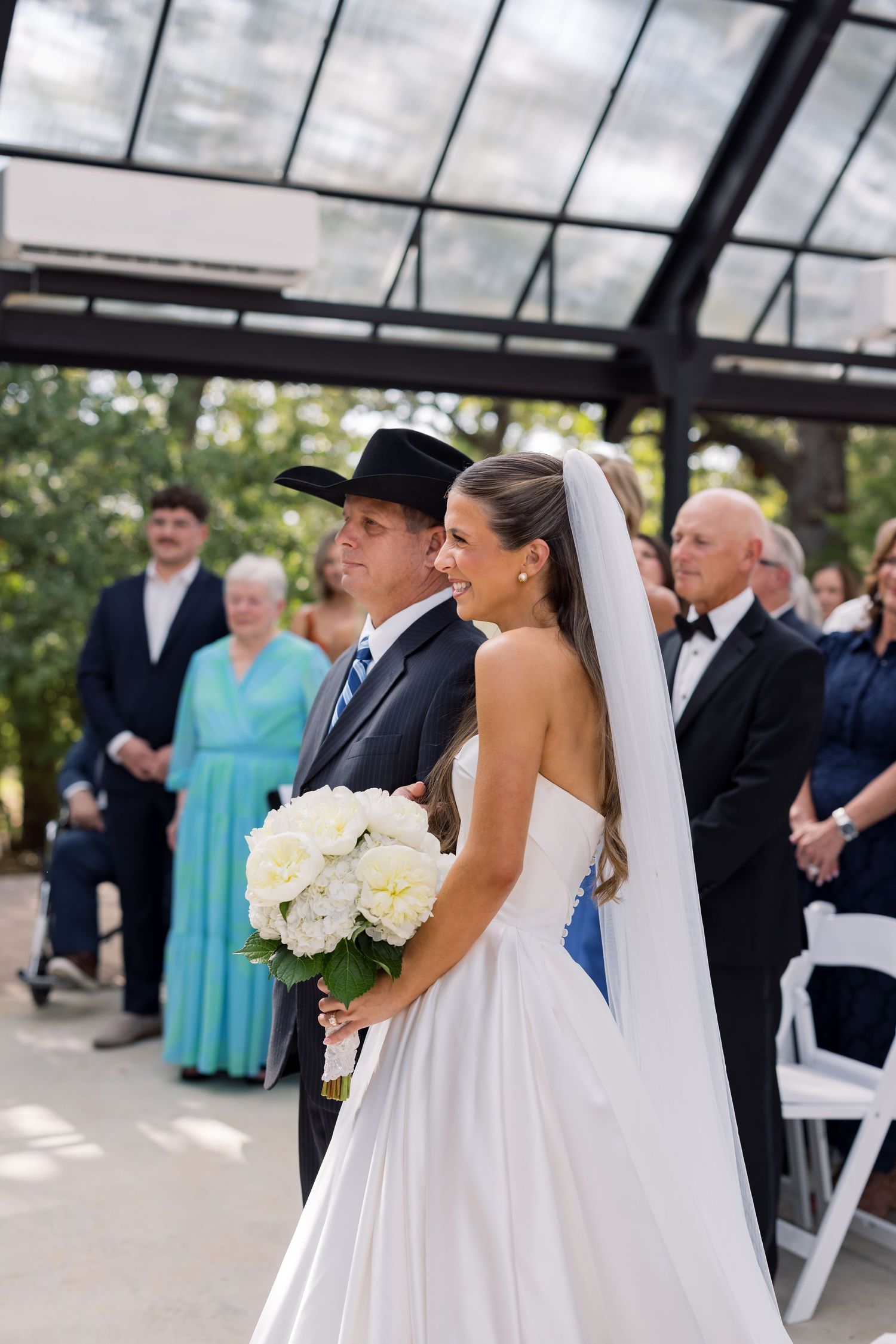 Bride in white strapless gown holding white bouquet walks with groom in black suit and cowboy hat at outdoor wedding ceremony.