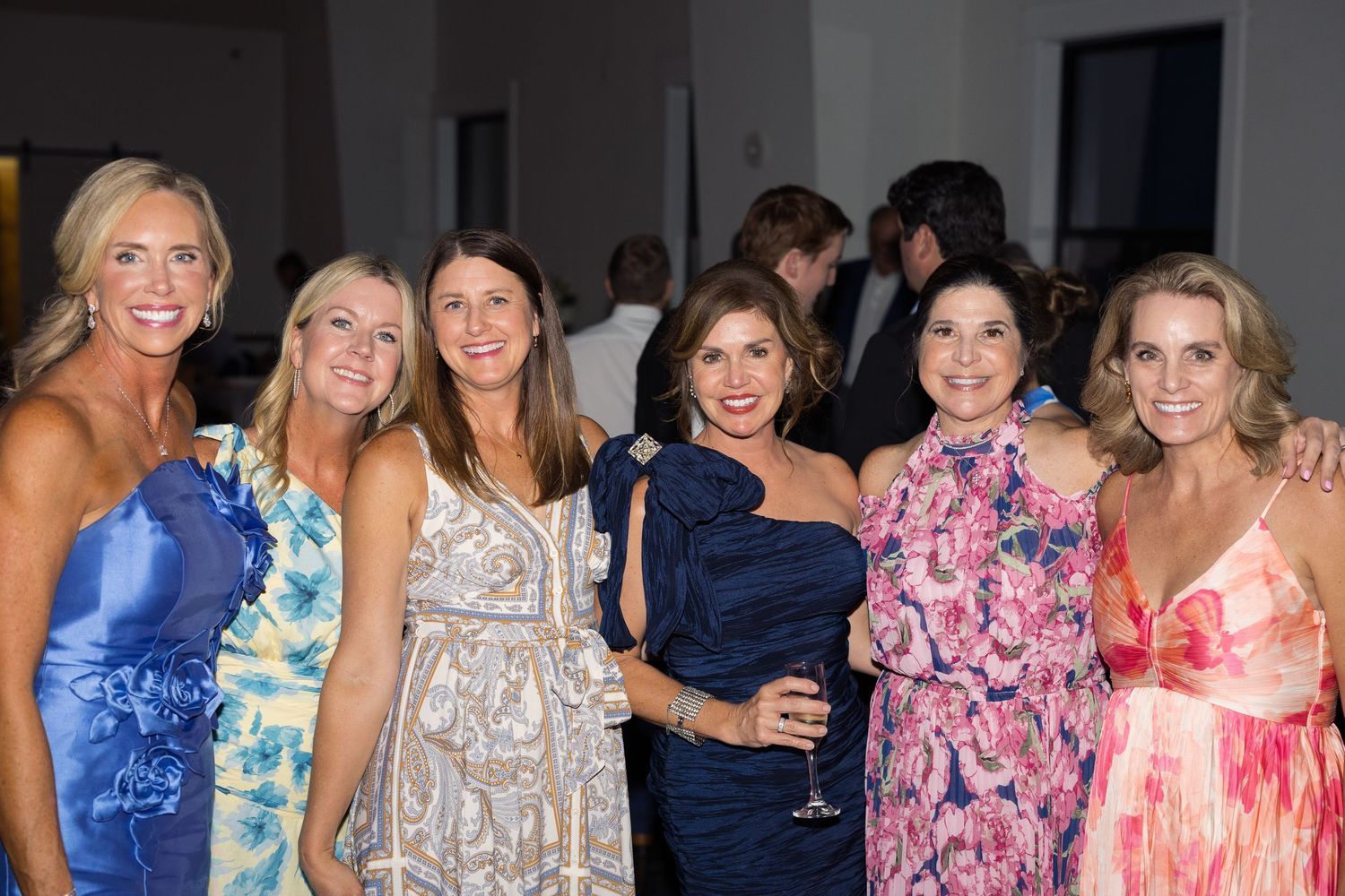 Six women in colorful dresses smile together at an elegant evening social event.