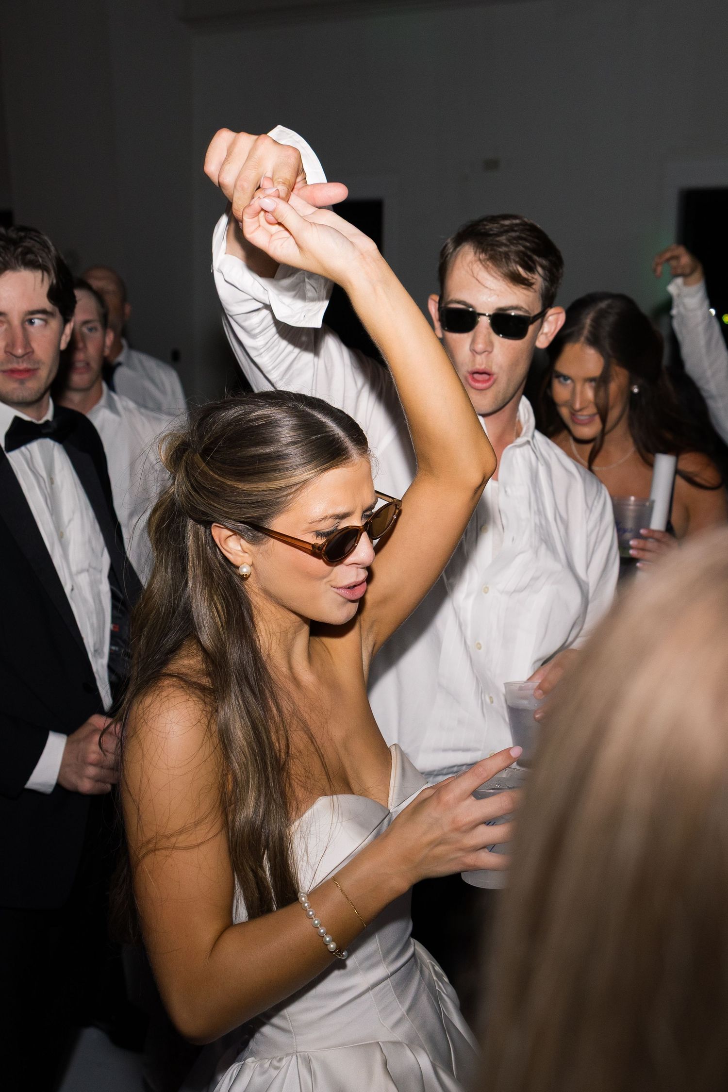Bride and groom dancing at wedding reception, both wearing sunglasses, surrounded by guests in formal attire.