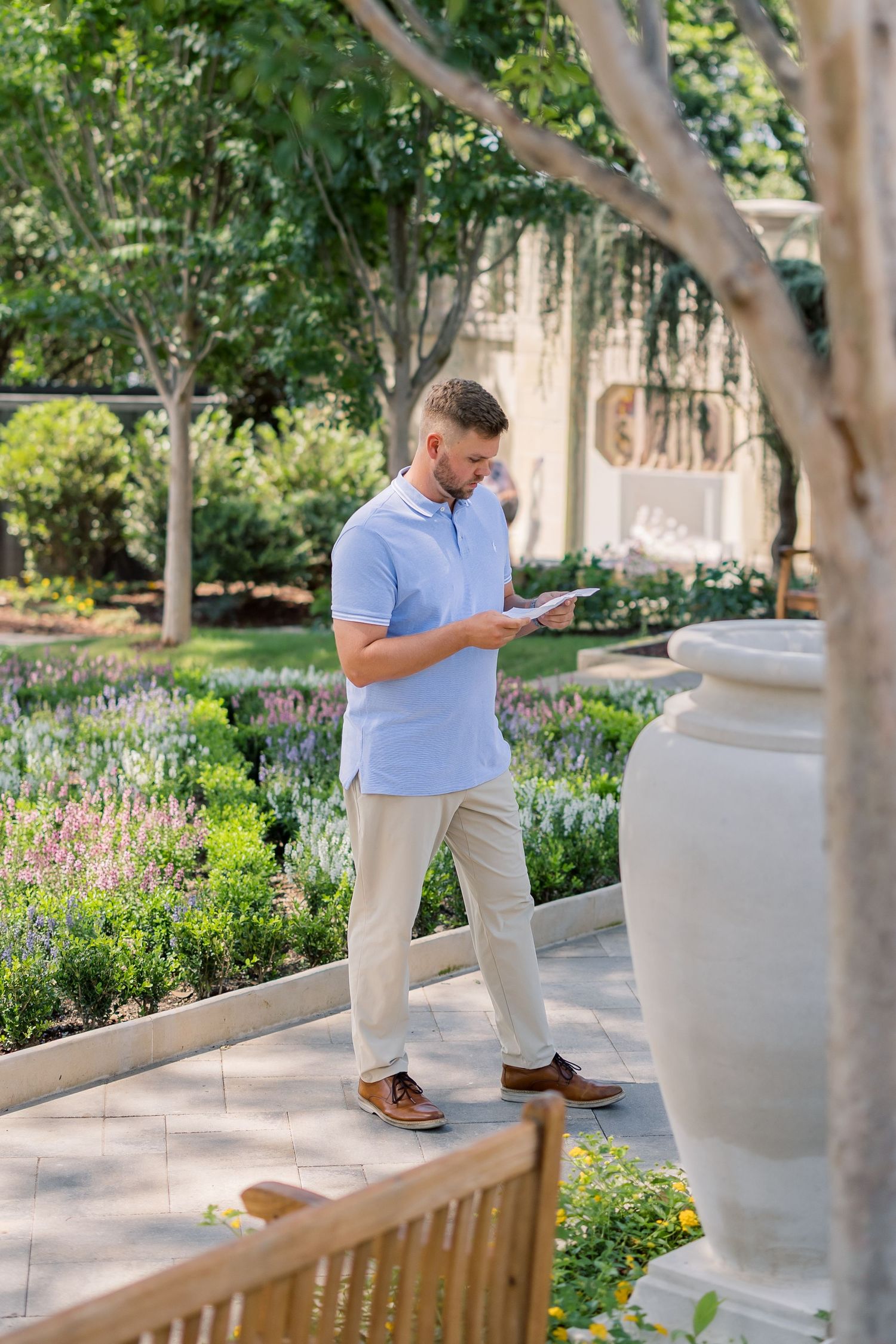 Man in blue polo shirt reading a document while walking through a lush garden with colorful flowers and trees.