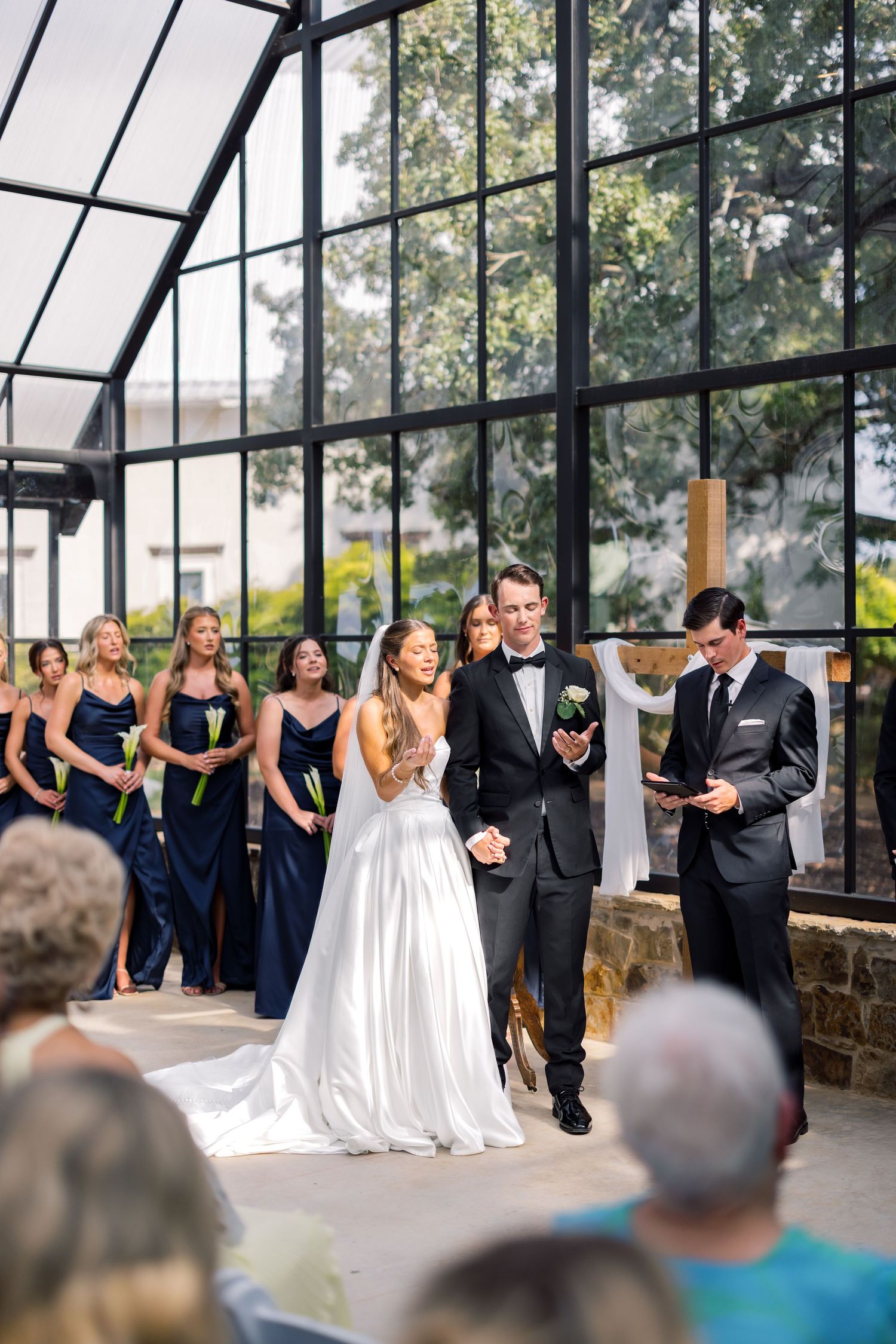 Bride and groom exchange vows in elegant glass chapel with bridesmaids in navy dresses during outdoor wedding ceremony.