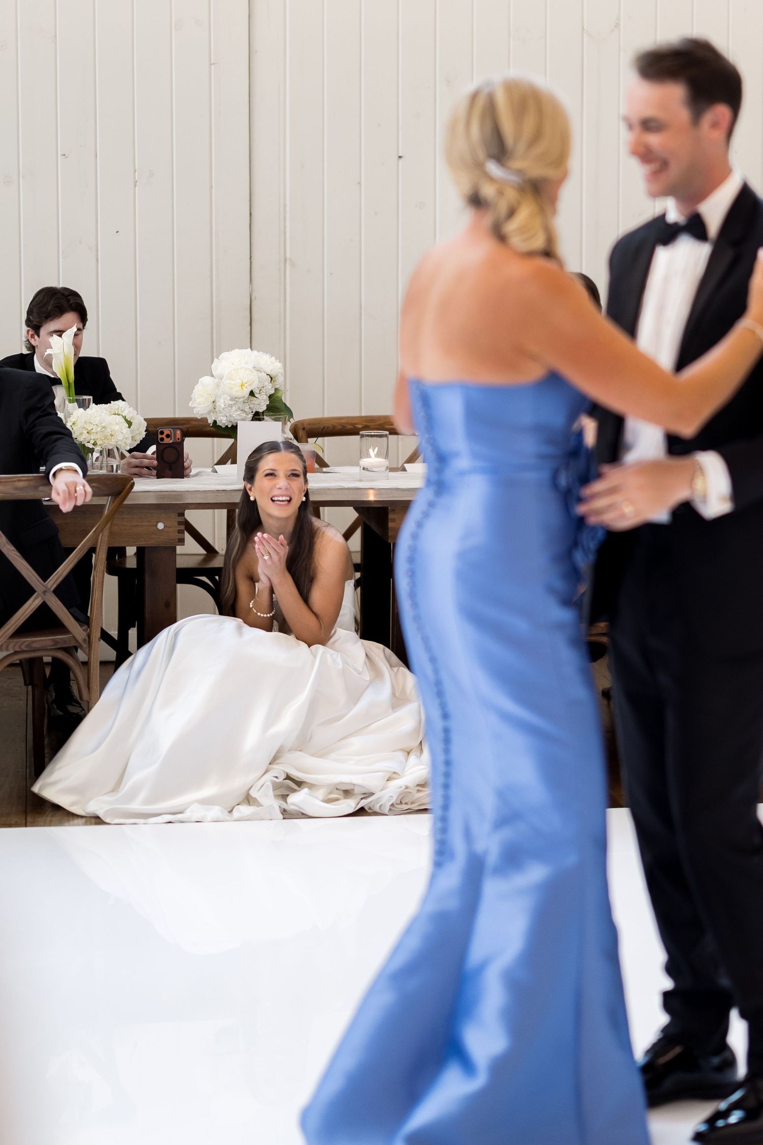 Bride clapping as groom dances with woman in blue gown at elegant wedding reception.