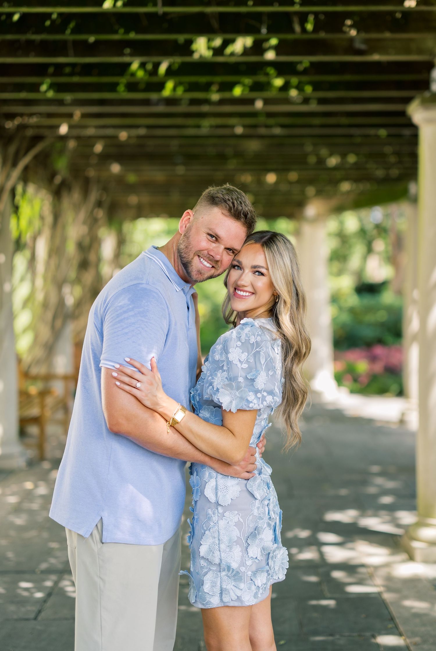 Couple embracing and smiling under a pergola in a lush garden, wearing coordinating light blue outfits.